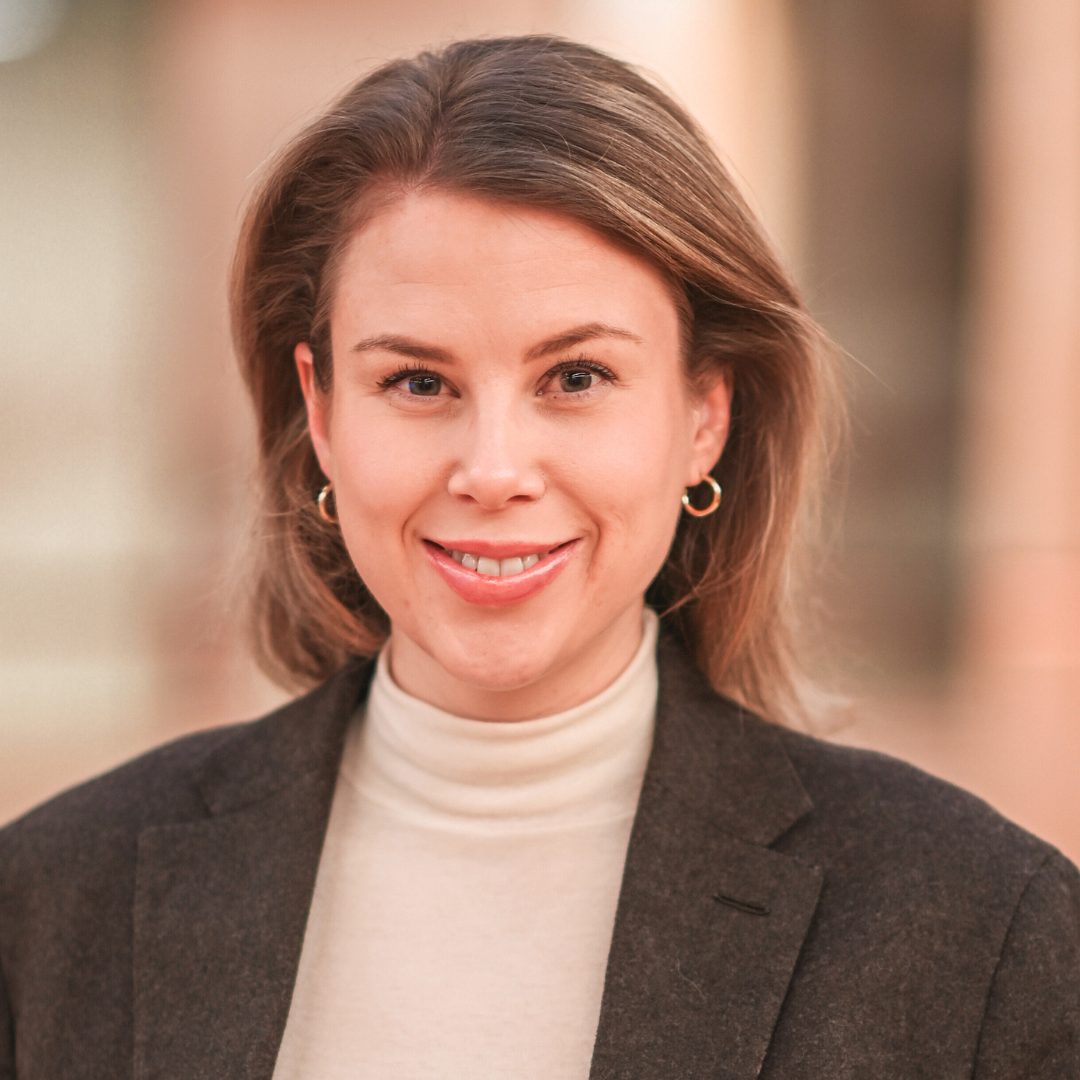 Woman with light brown hair smiles, wearing a cream turtleneck and dark blazer.