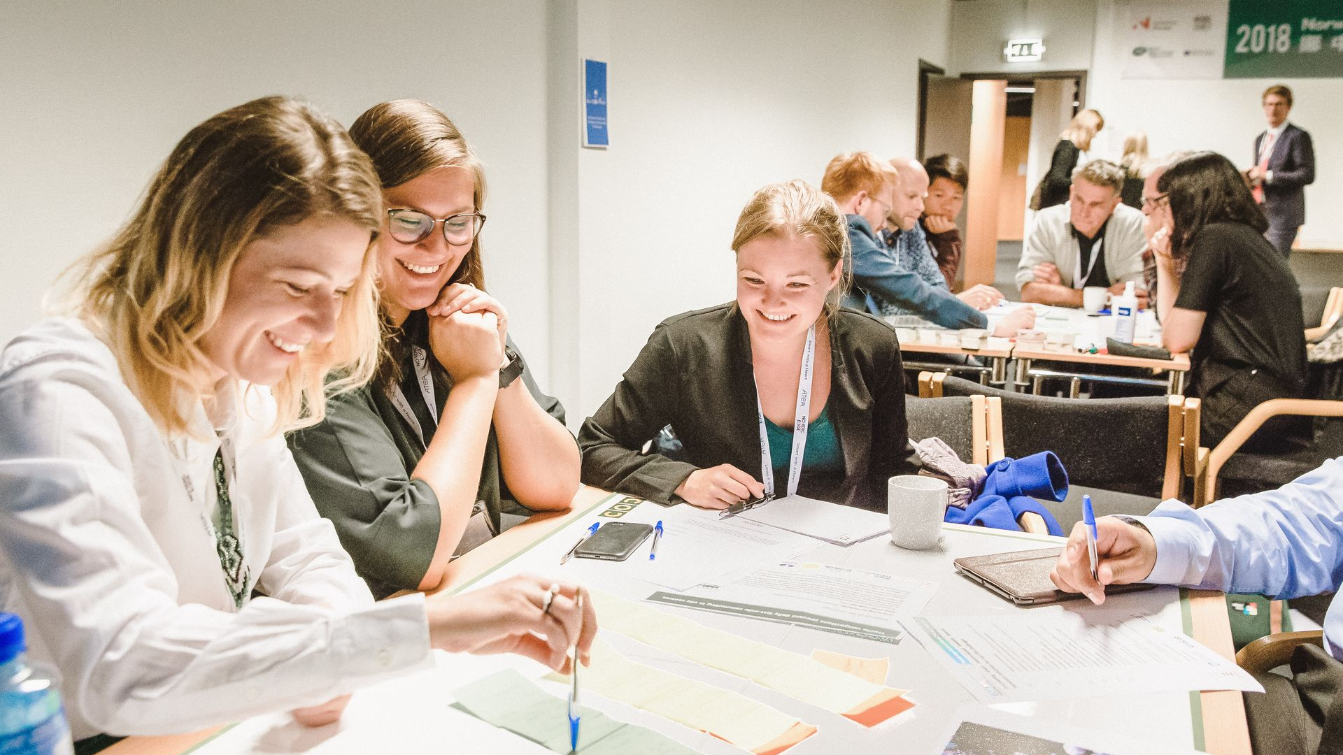 People in business attire at a table, discussing. Woman in white sweater looks up; German flag present.