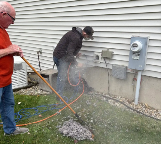 Person working on exterior electrical, next to a house. Another person sweeps debris from the lawn.