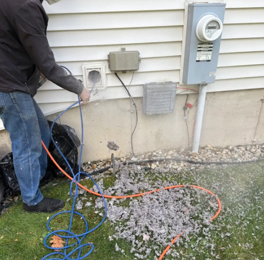 Person cleaning a dryer vent, blowing lint onto the grass outside a house.