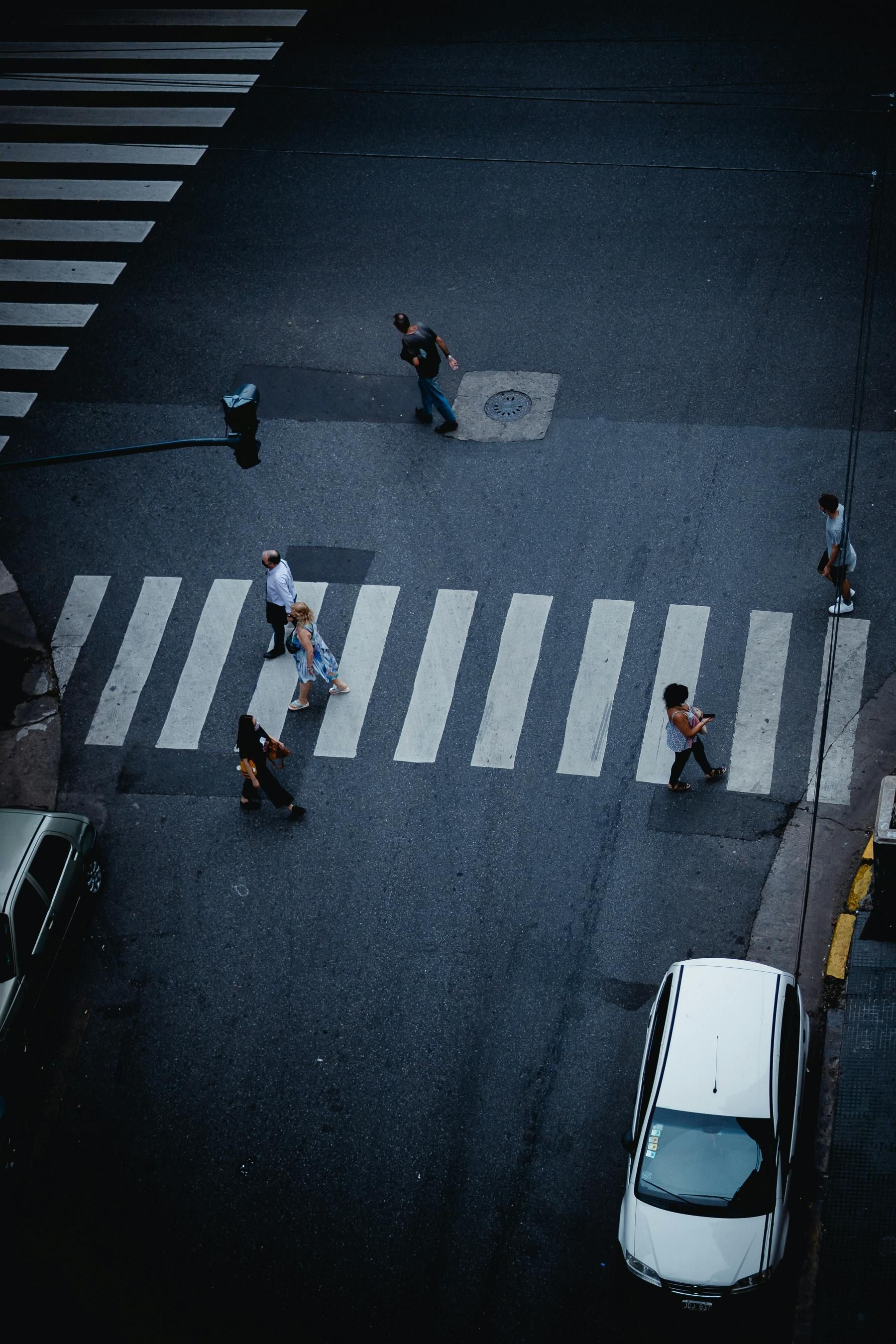 People crossing a street at a crosswalk. Dark asphalt with white stripes. A white car is parked nearby.