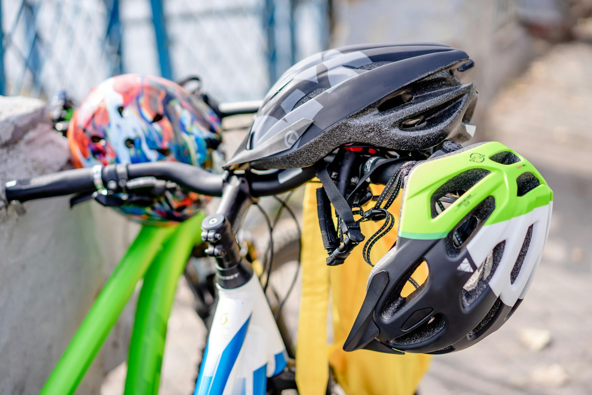 Three bike helmets hanging on a bicycle's handlebars; one black, one lime green, and one colorful.