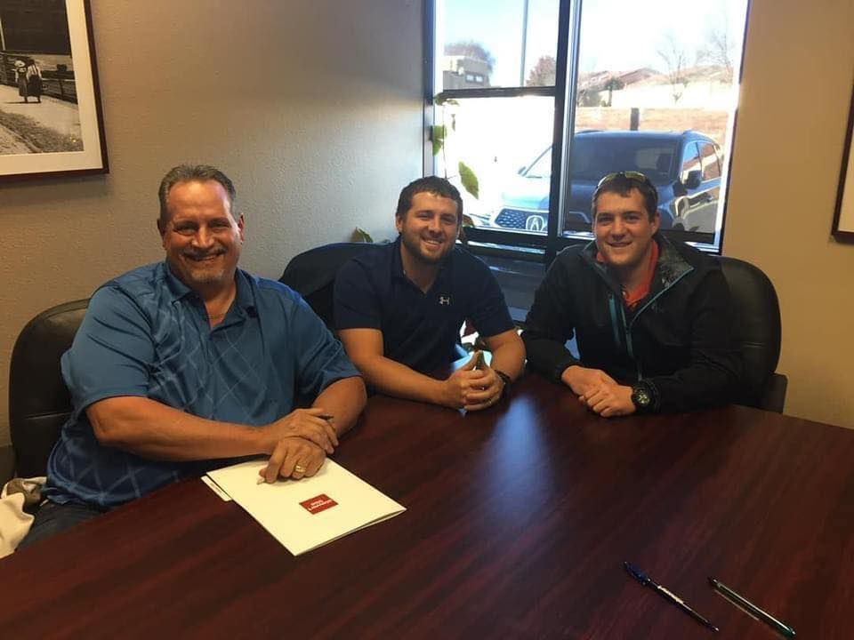 Three men seated at a table, smiling. A document with logo on table. Office setting with window.