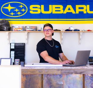 A man sitting at a desk with a laptop in front of a subaru banner