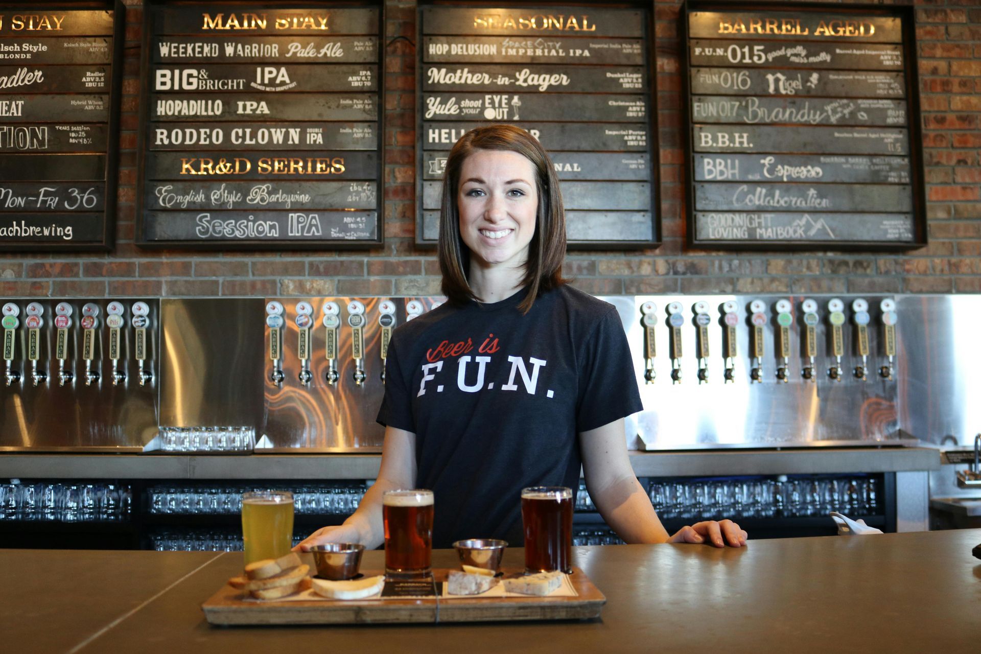 A woman wearing a shirt that says fun is standing at a bar holding a tray of beer.