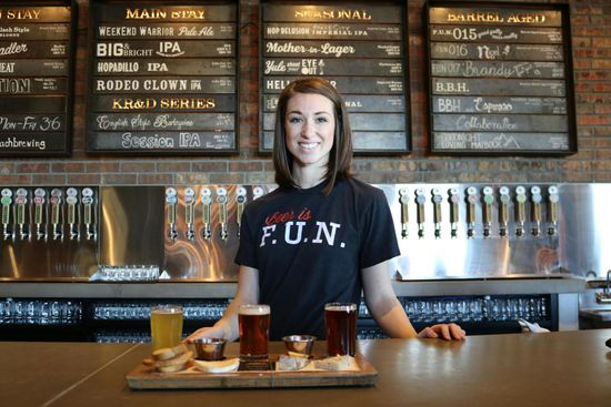 A woman wearing a shirt that says fun is standing at a bar holding a tray of beer.