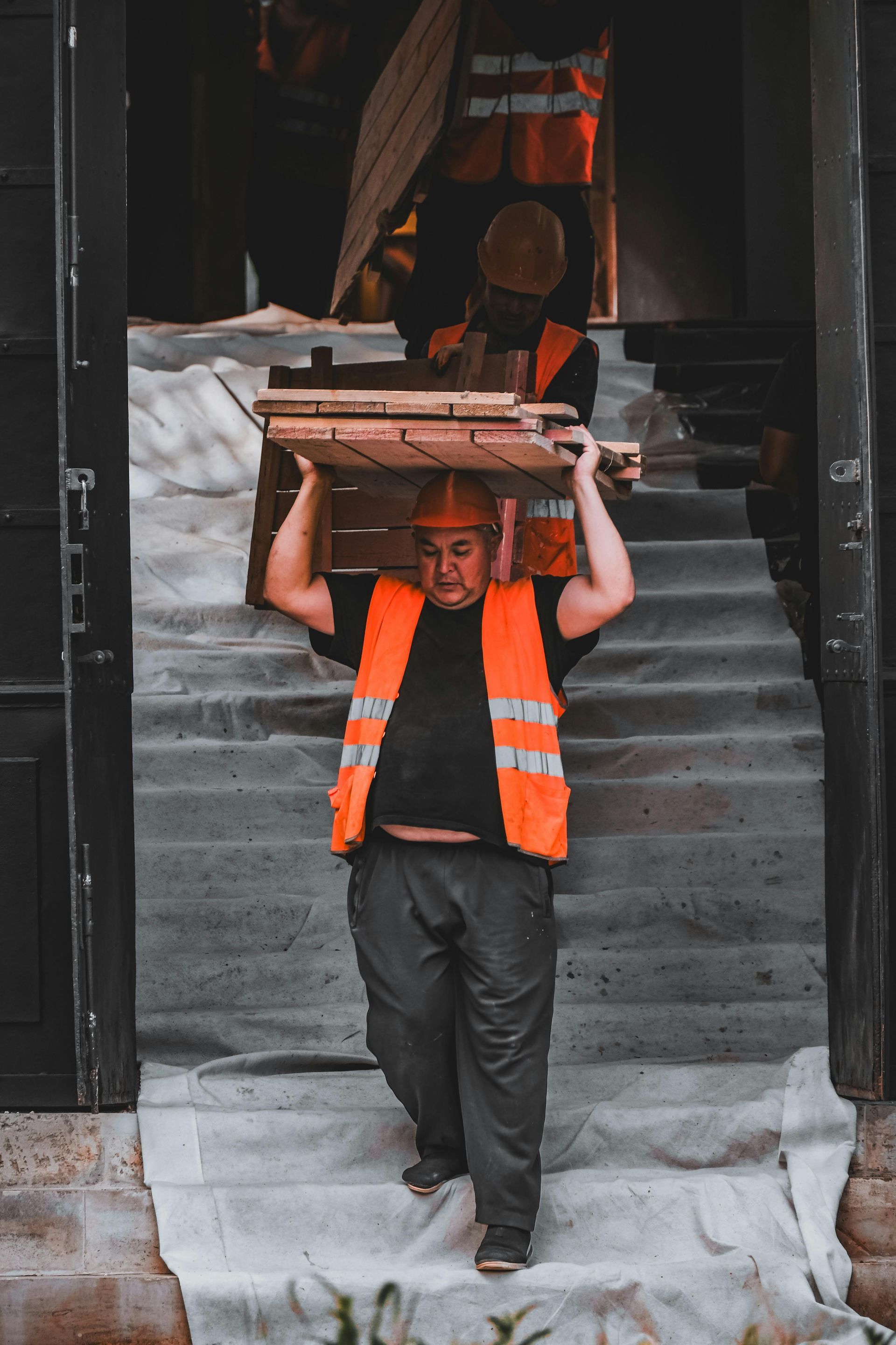 A construction worker in a high-visibility orange vest carries a wooden crate down a staircase draped in a white cloth.