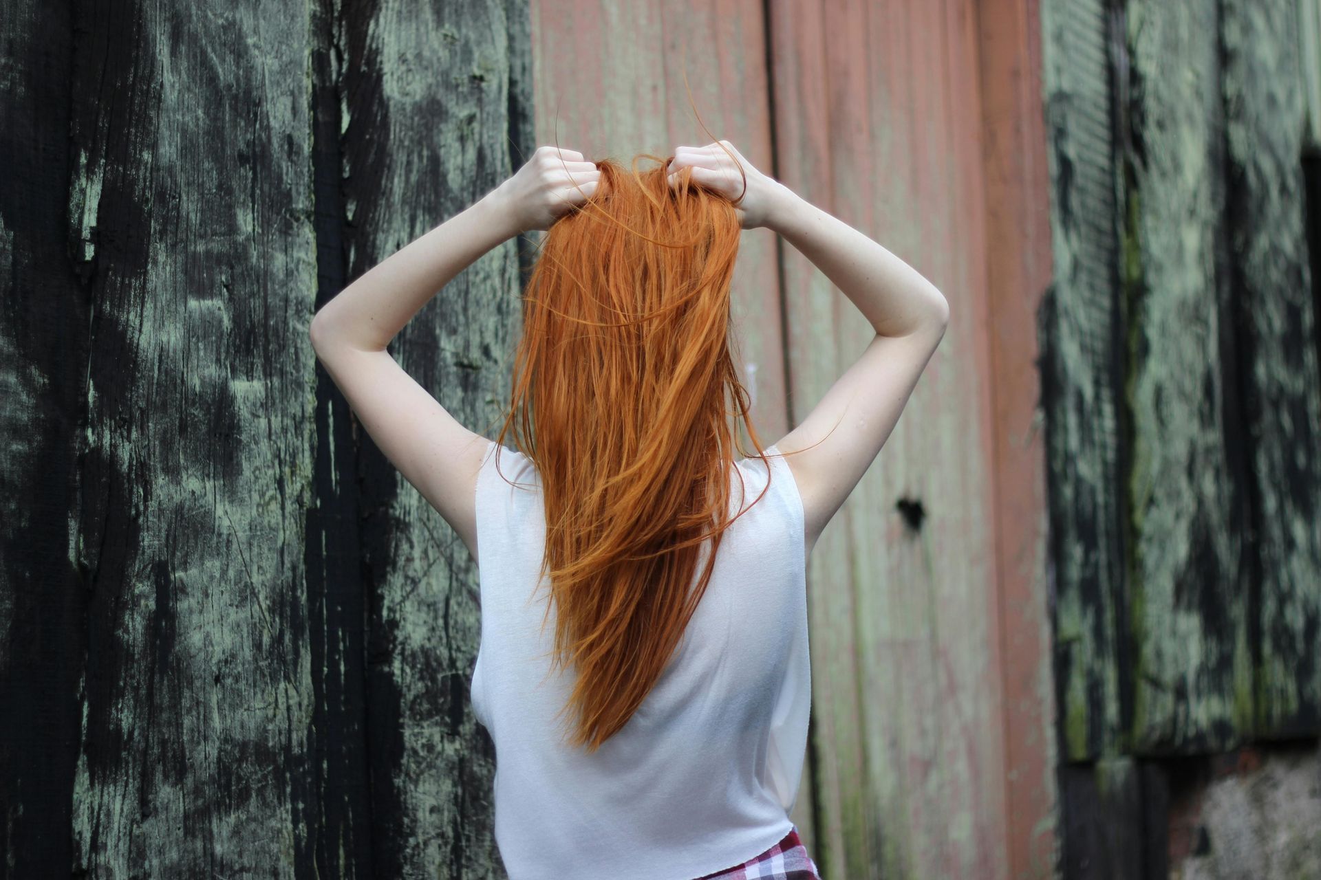 A person with long, vibrant red hair seen from behind, holding their hair up with both hands against a weathered wall.