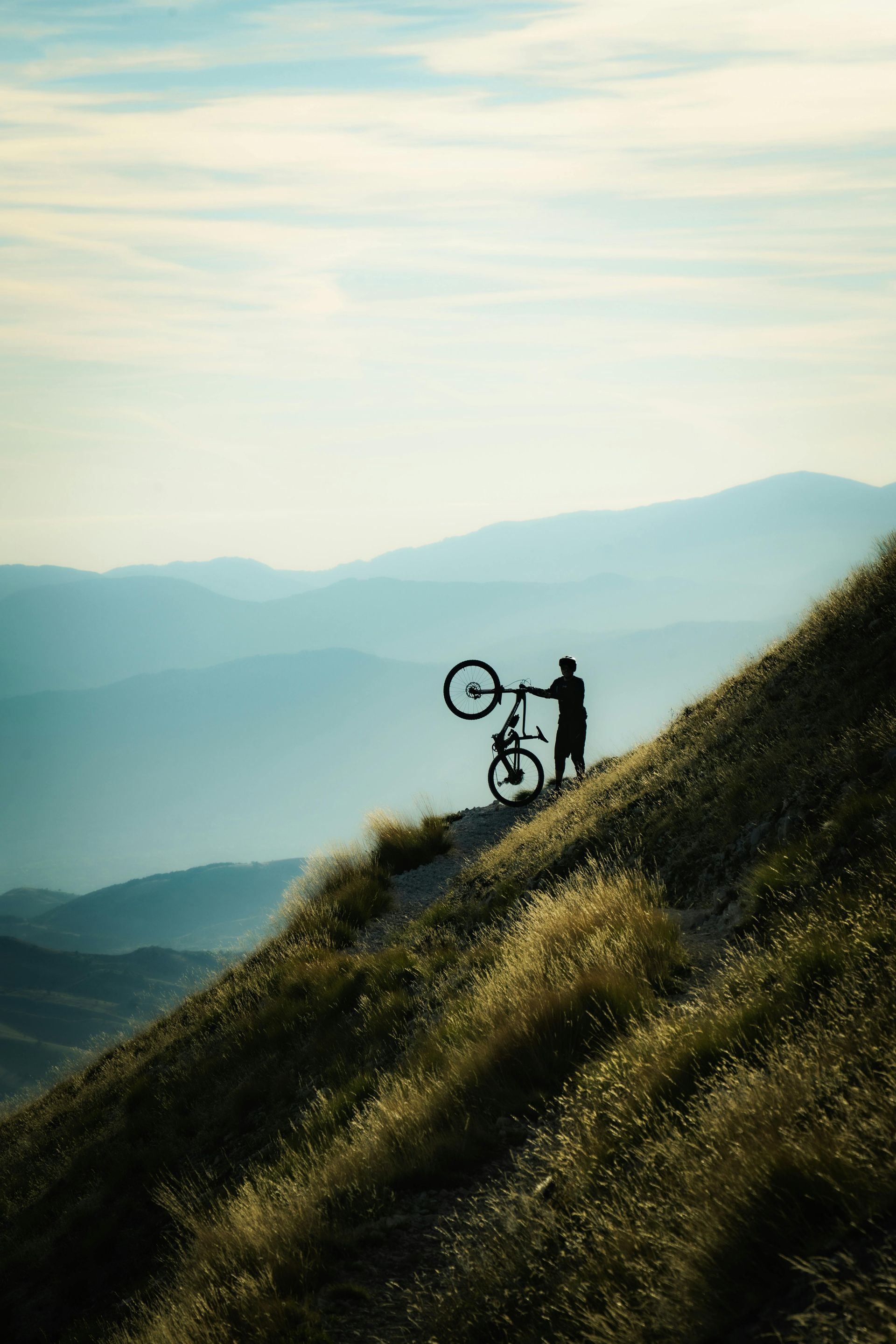 Silhouette of a person holding a mountain bike on a grassy mountain ridge, overlooking layered blue-toned mountains.