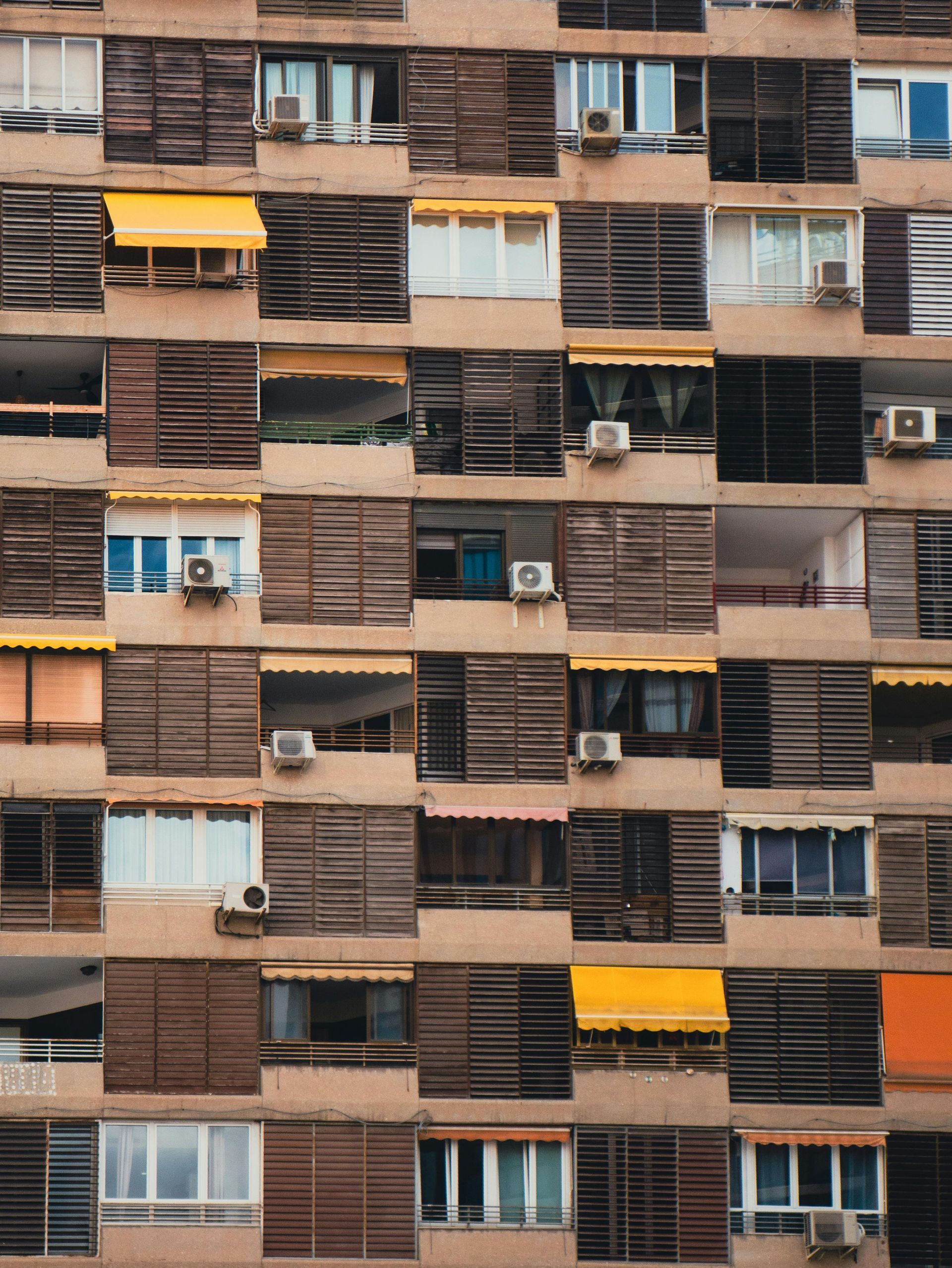 Apartment building facade with wooden panels, air conditioners, and a few yellow awnings.