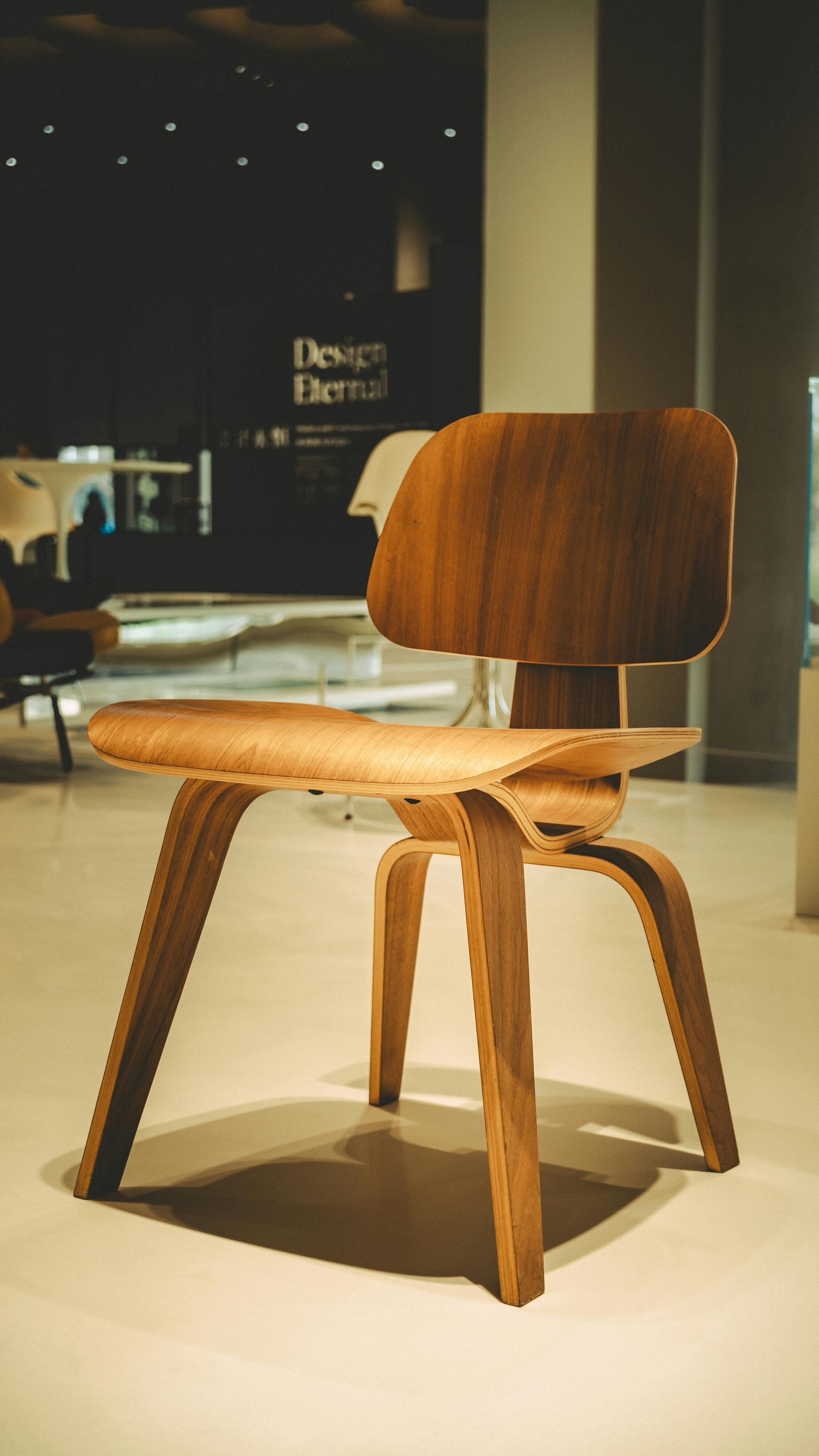 A light-colored plywood chair with a mid-century modern aesthetic displayed in a gallery setting.