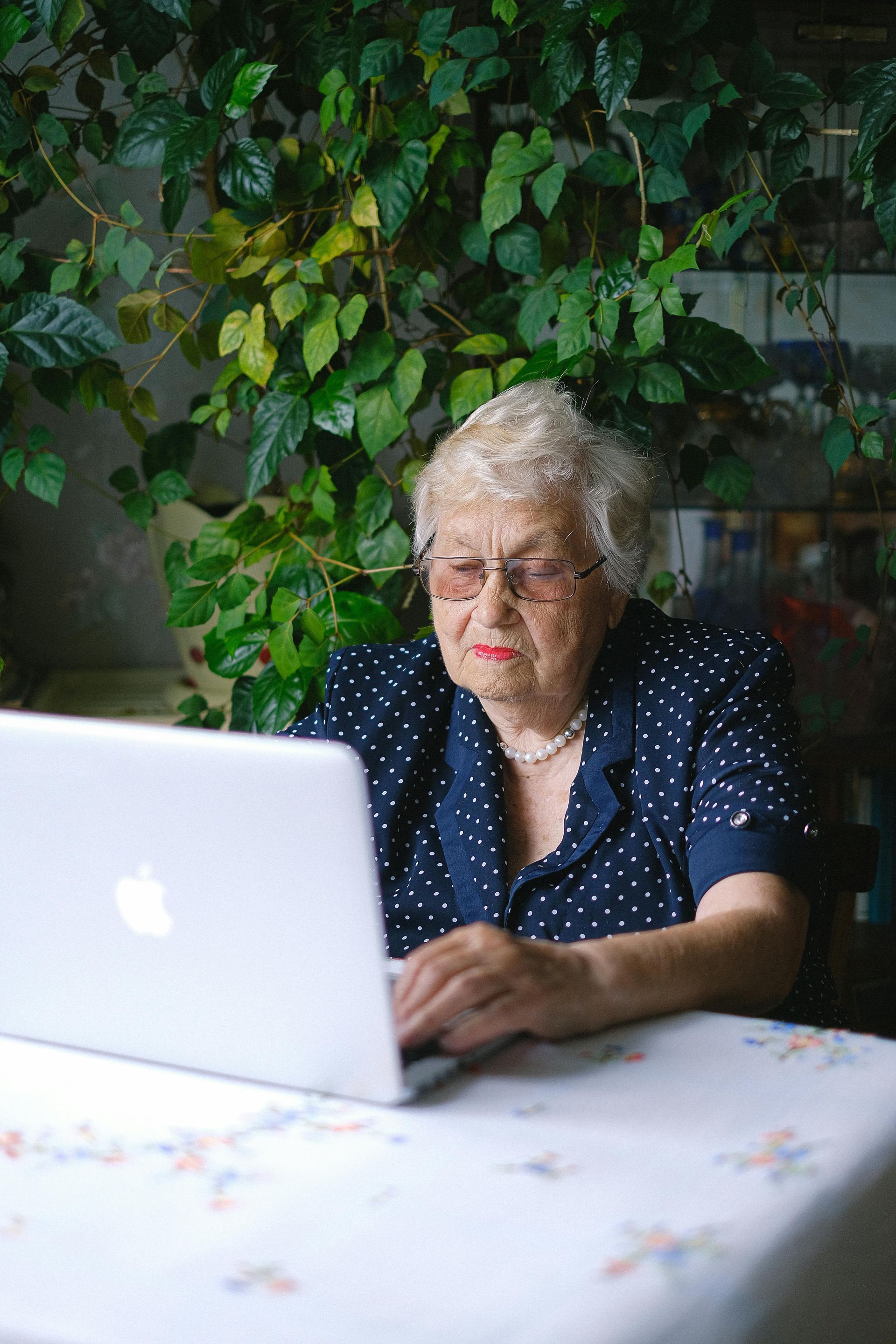 An elderly woman is sitting at a table using a laptop computer.