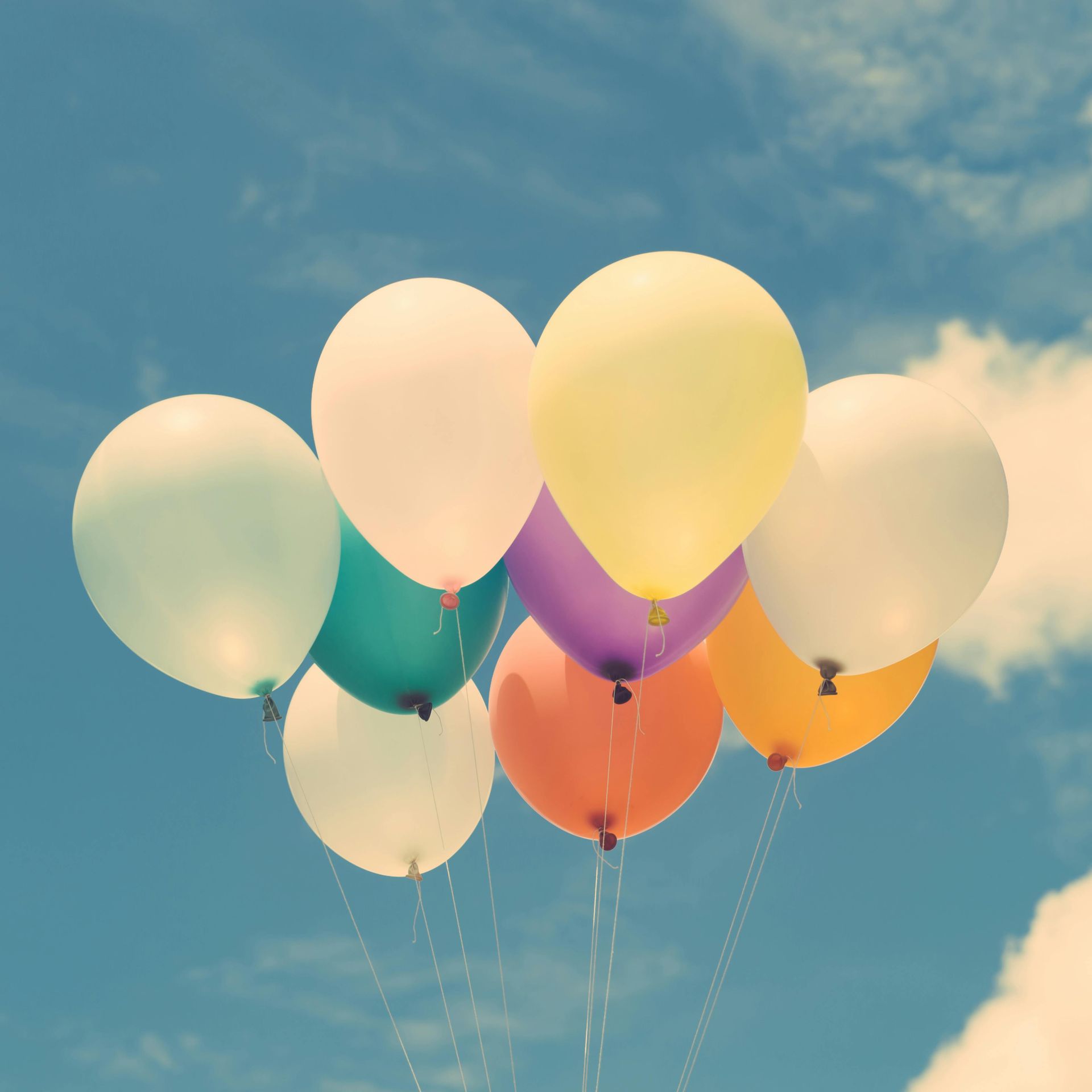 A bunch of colorful, pastel-toned balloons floating against a blue, cloudy sky.