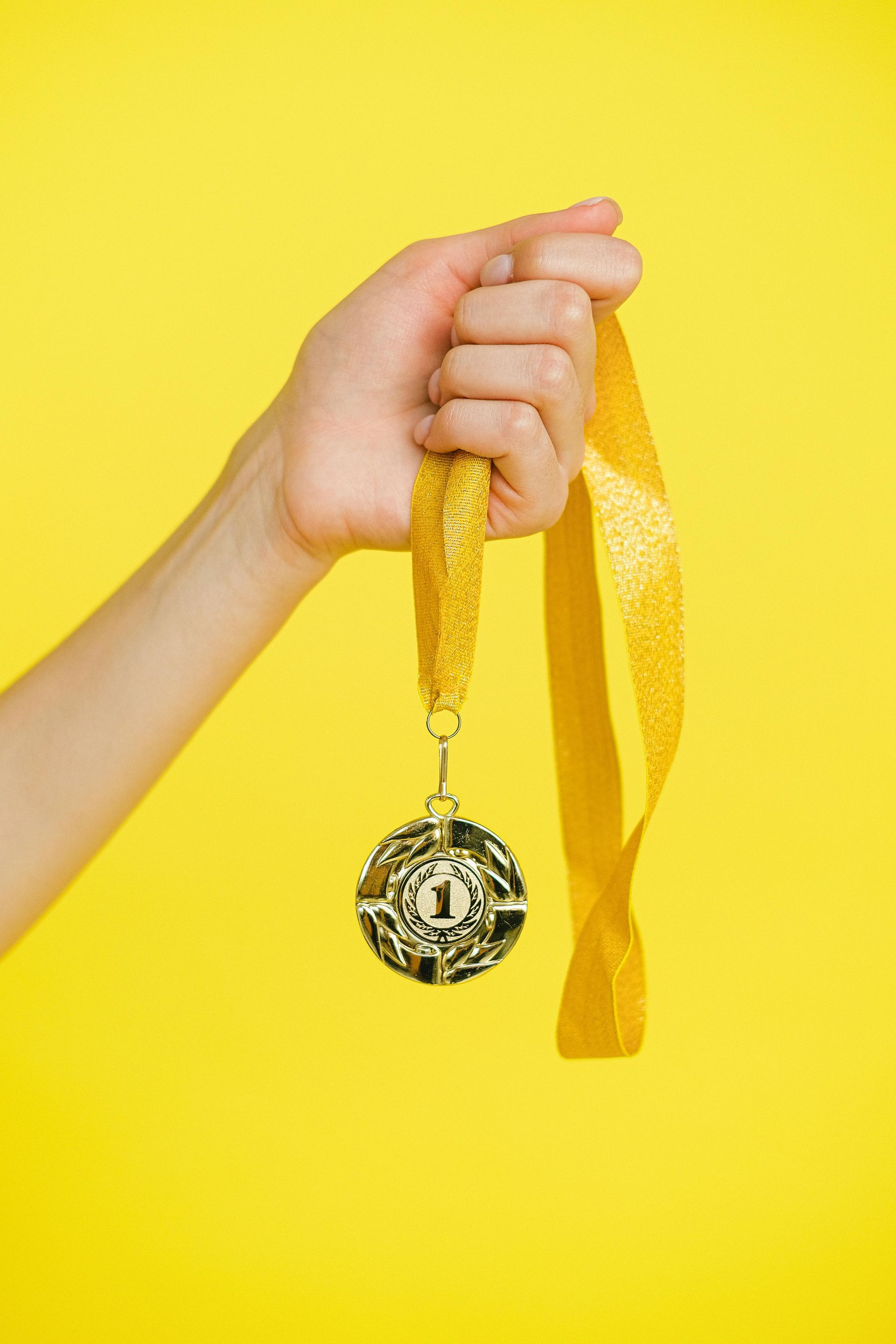A hand holds up a gold medal with the number one on it against a solid yellow background.