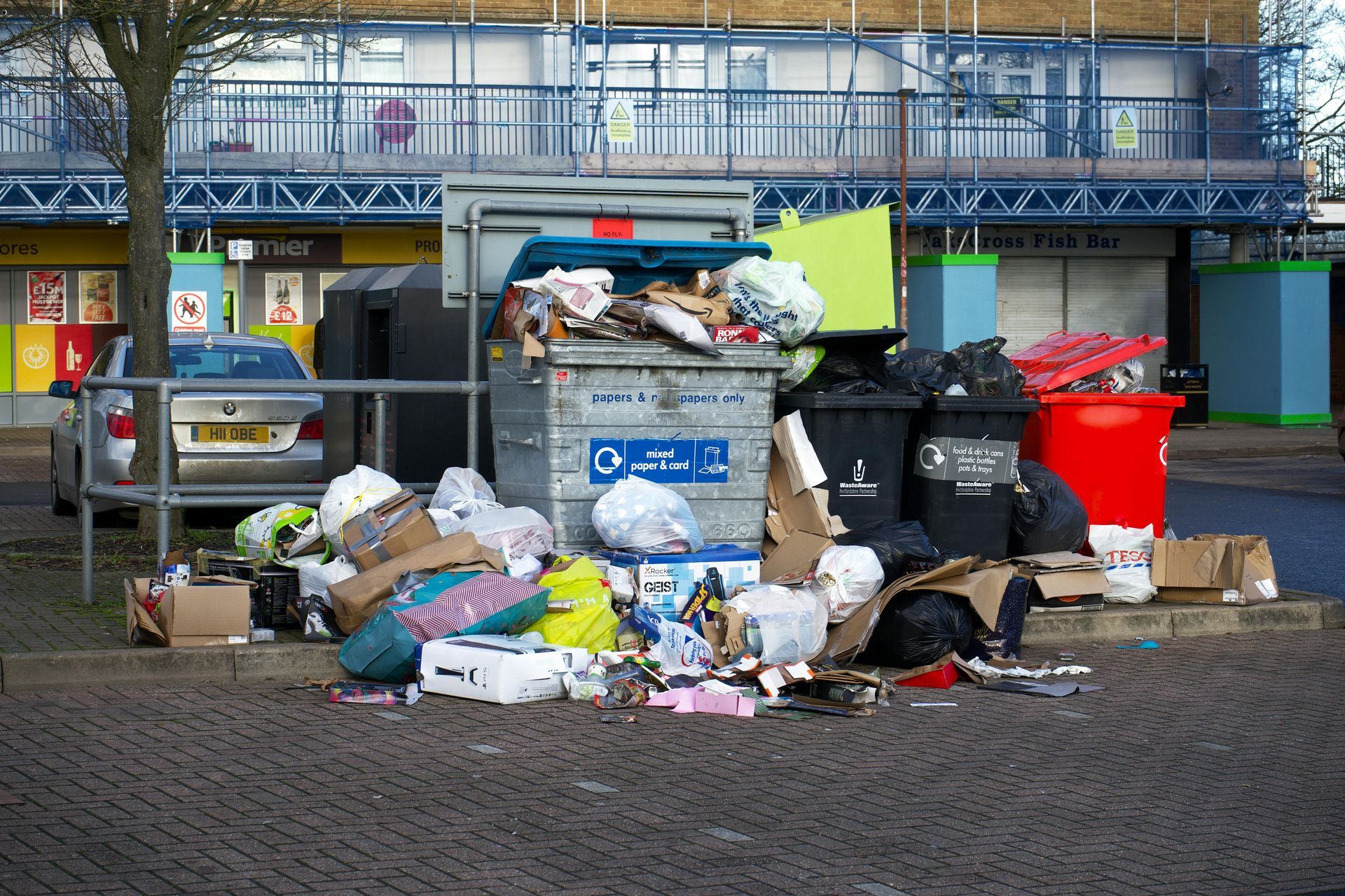 Overflowing metal and plastic dumpsters surrounded by loose garbage and cardboard boxes in a parking lot.