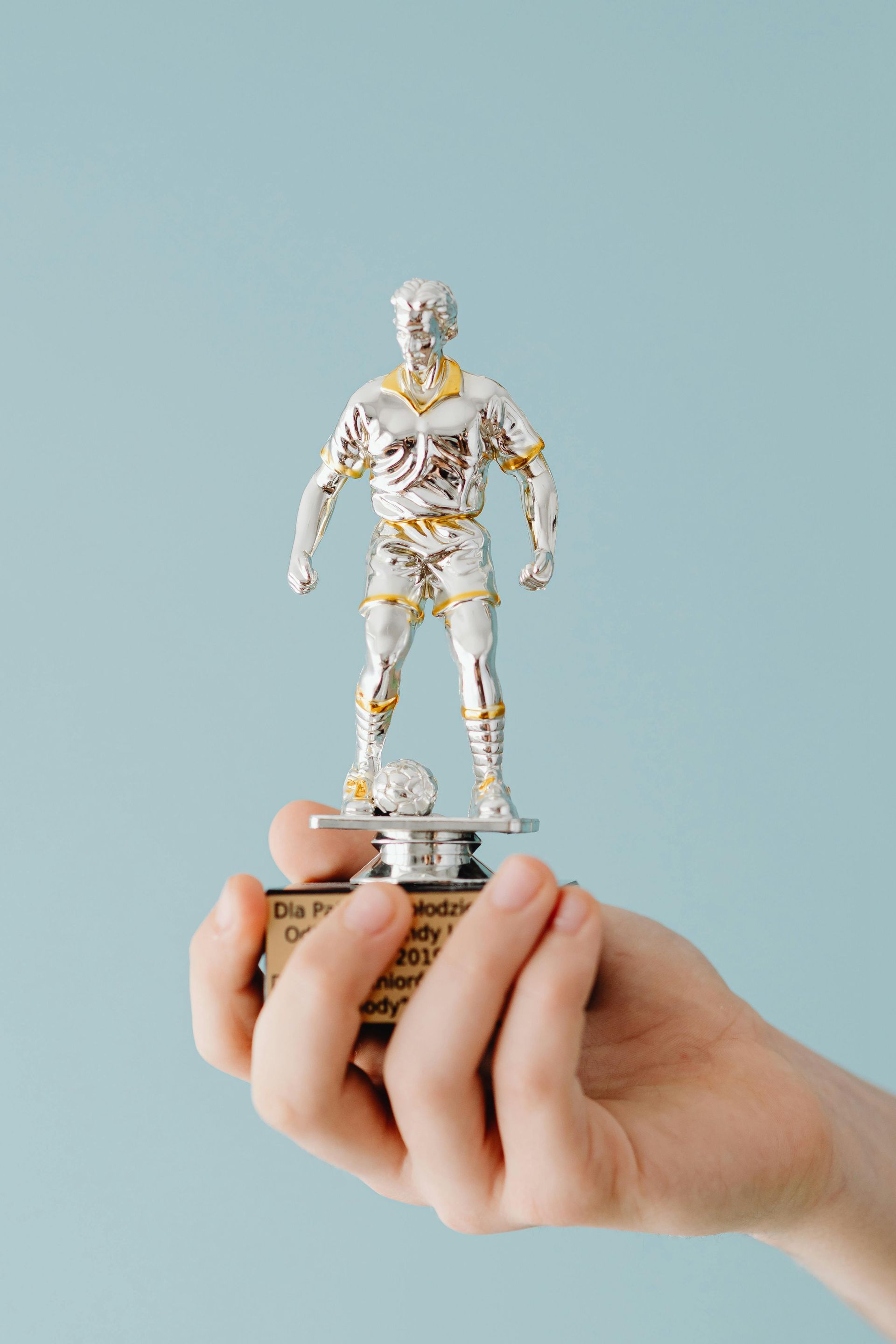 A hand holds a small silver soccer trophy with gold accents against a light blue background.