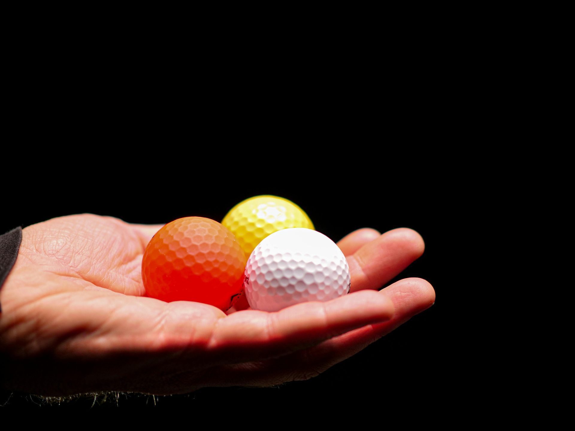 A person's open hand holds an orange, a yellow, and a white golf ball against a black background.