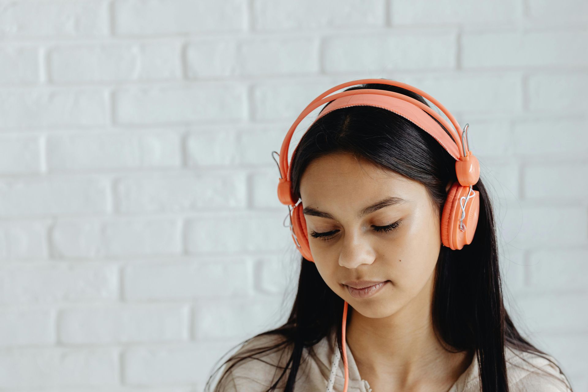 A person with long dark hair wears peach-colored over-ear headphones in front of a white brick wall.
