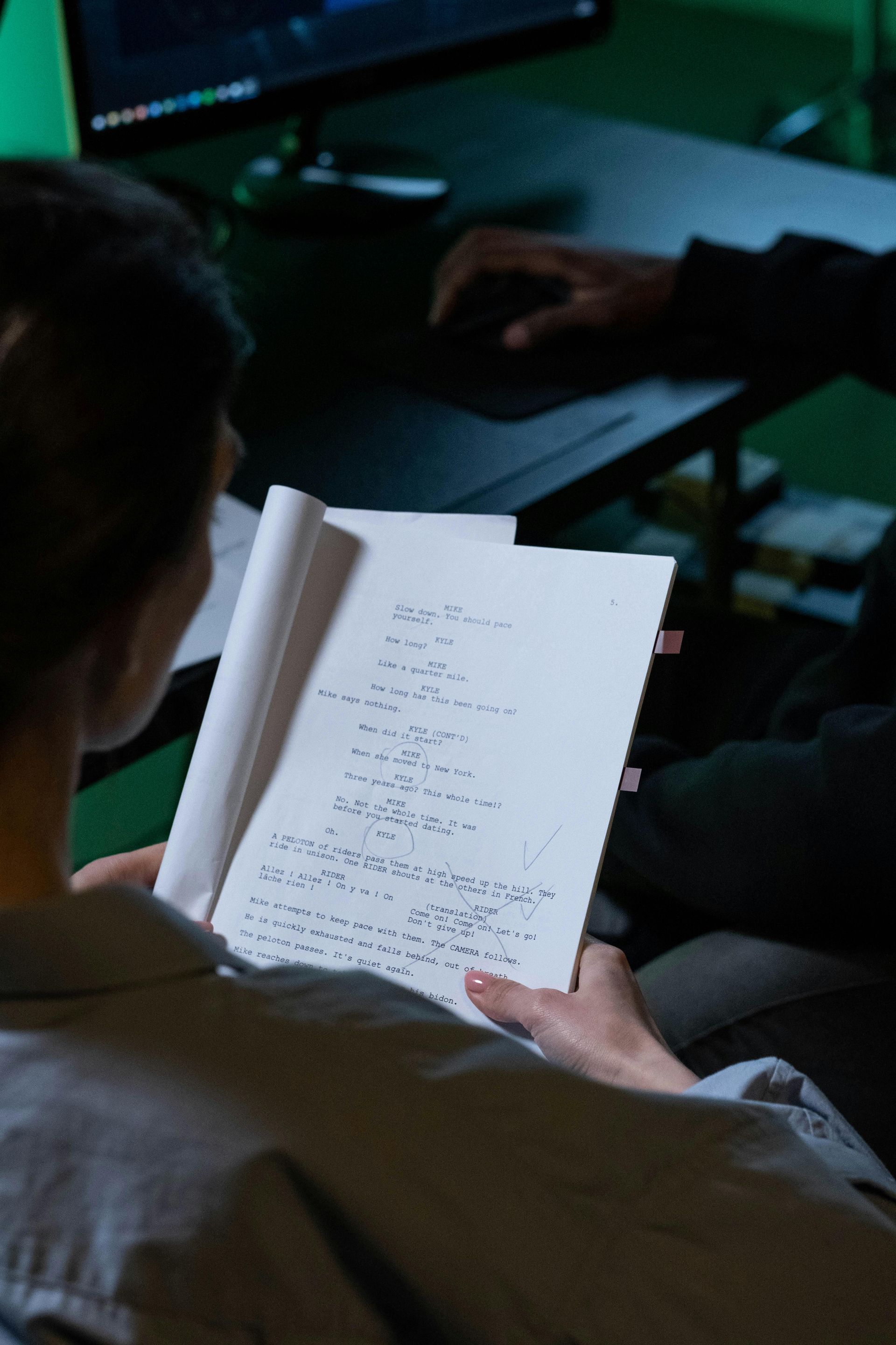 A person holds a handwritten document while another person works at a computer in a dimly lit, green-tinted office space.