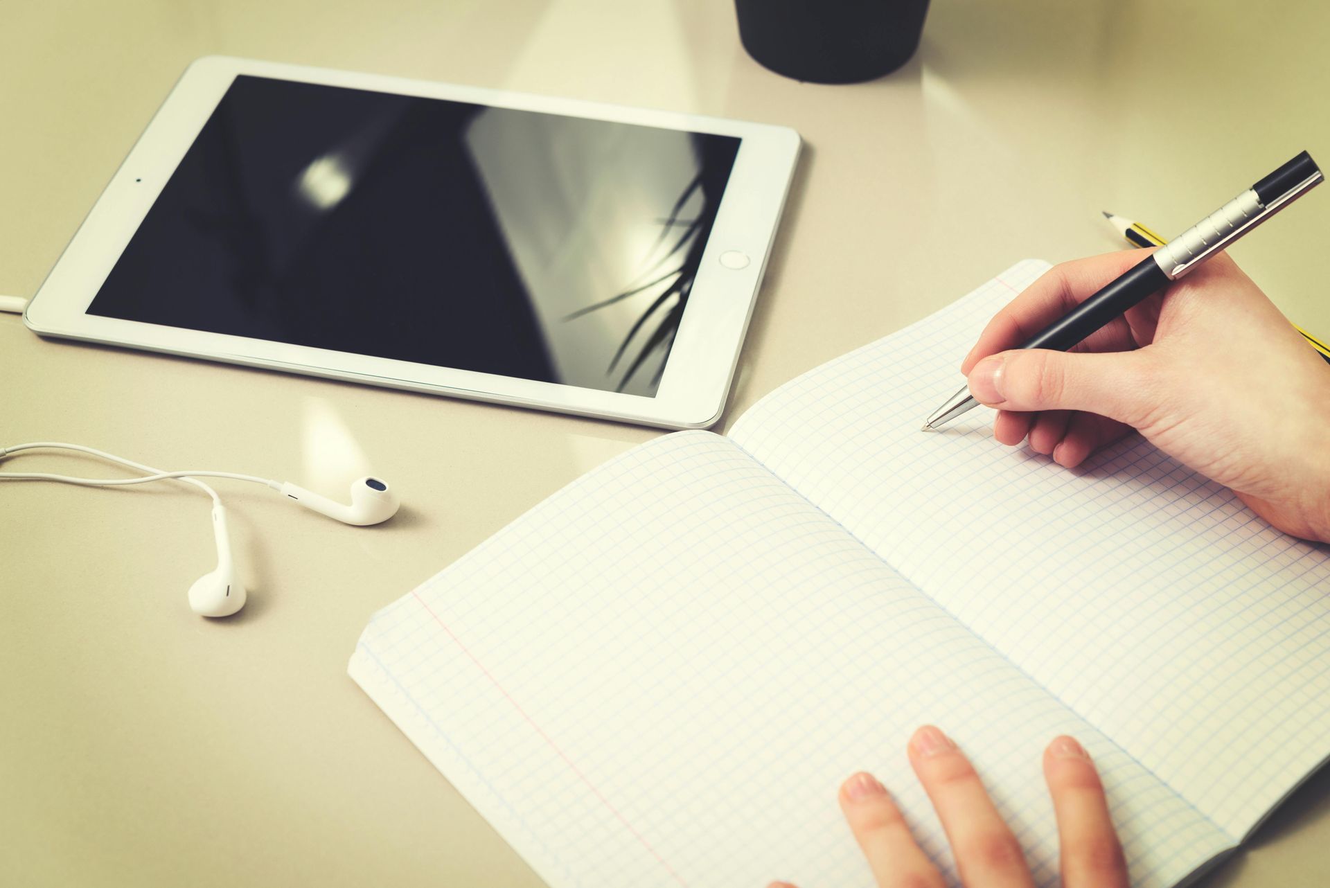 A person is writing in a notebook next to a tablet.