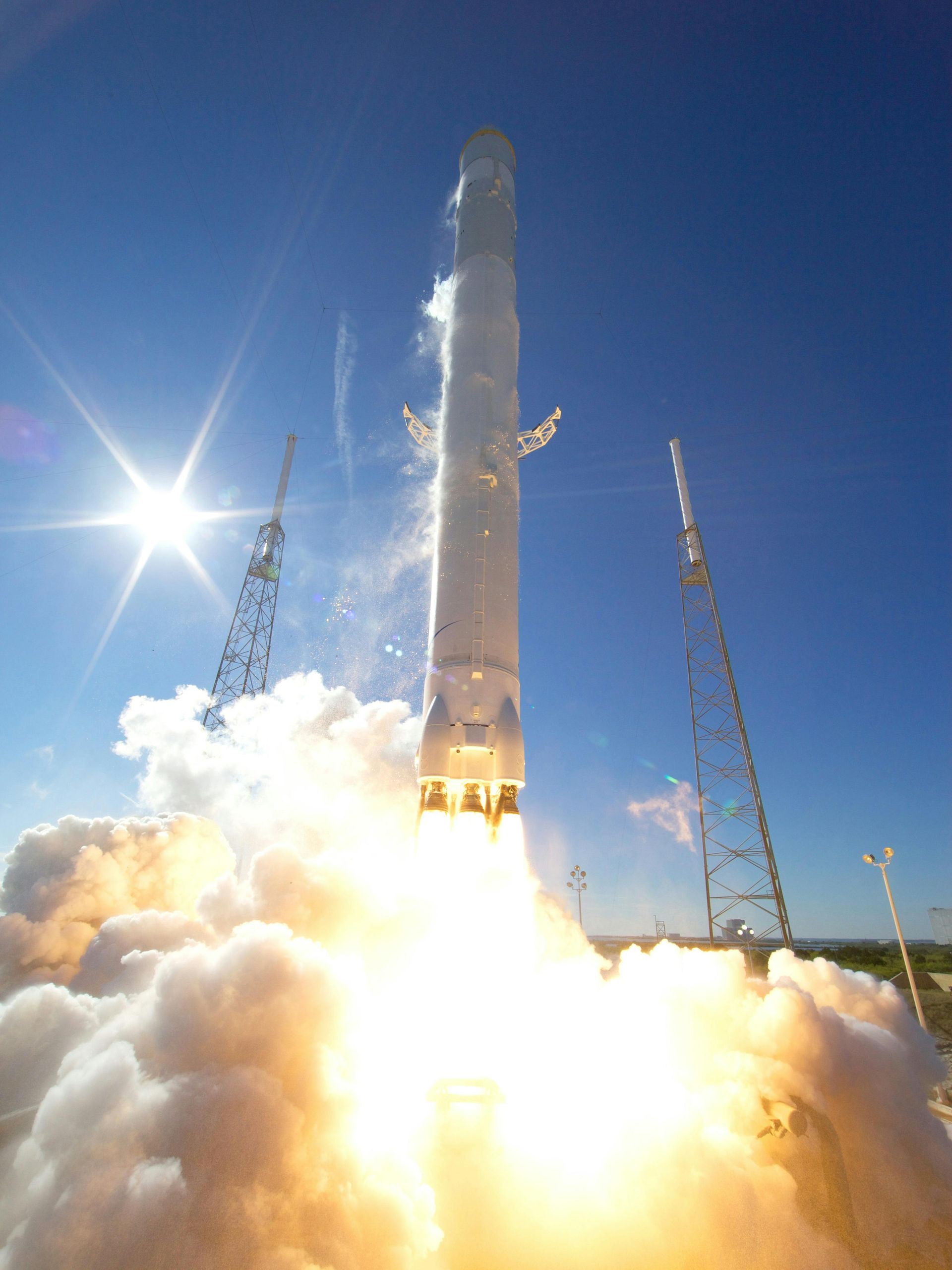 A white rocket launches into a clear blue sky, surrounded by bright fire and thick white smoke near two metal towers.