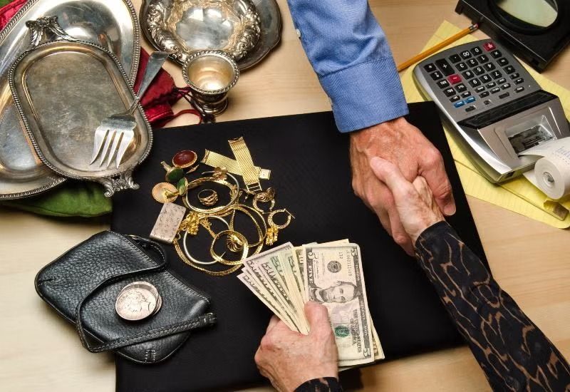 Hands counting cash beside jewelry, silverware, and a calculator on a table