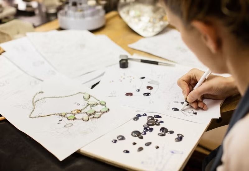 Person sketching jewelry designs on paper at a desk with beads and tools