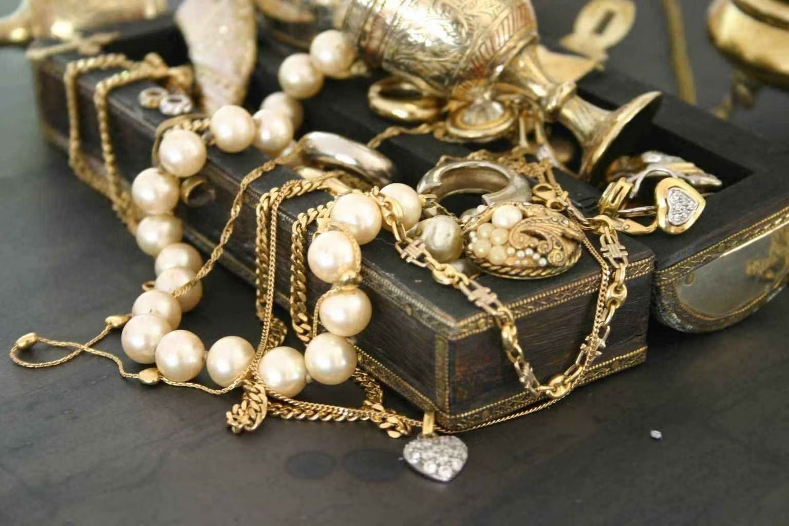 Gold jewelry and pearl necklace arranged on a black box, with a decorative key and watch.