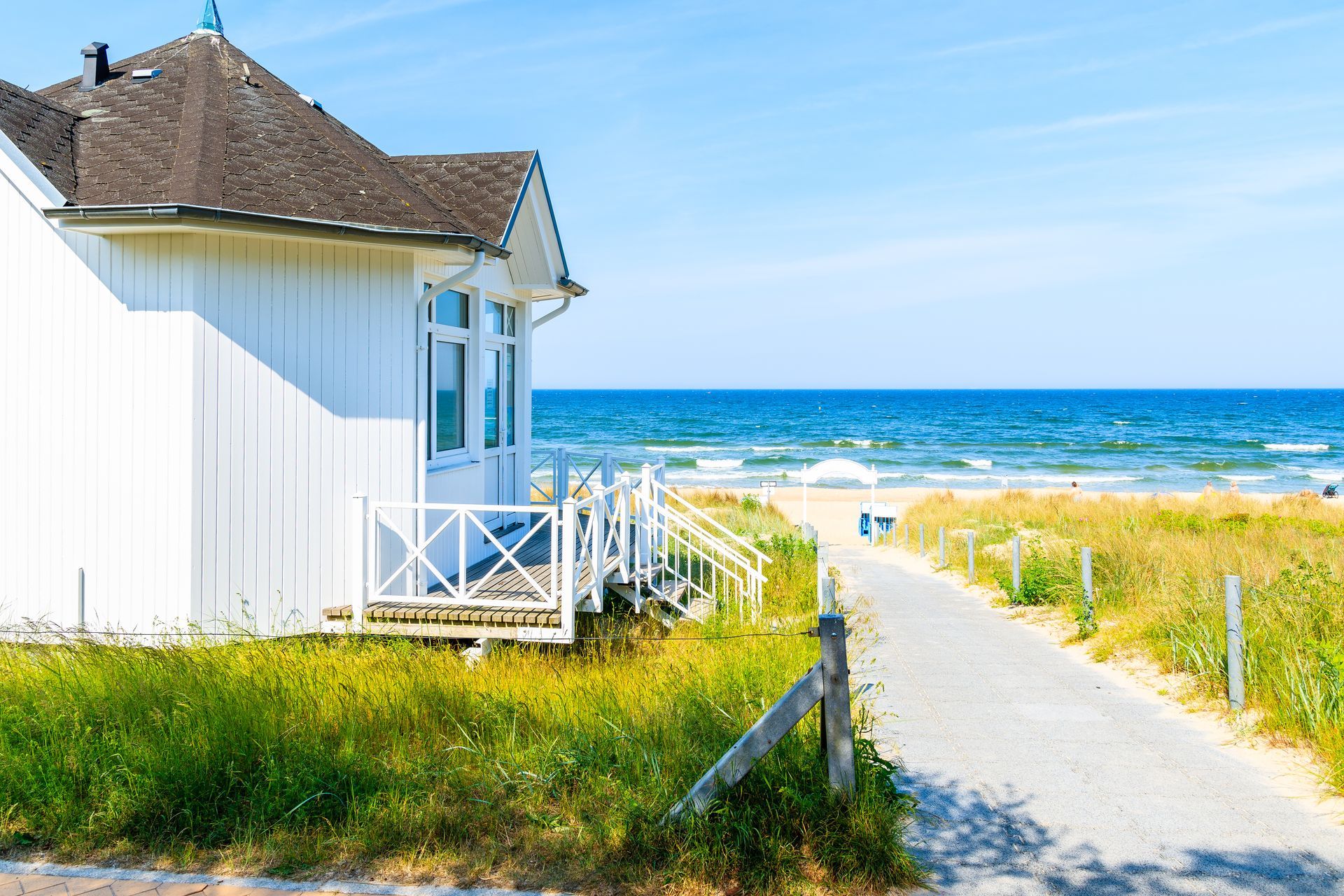 A white cottage with a dark roof stands beside a sandy path leading to a blue ocean under a clear sky.