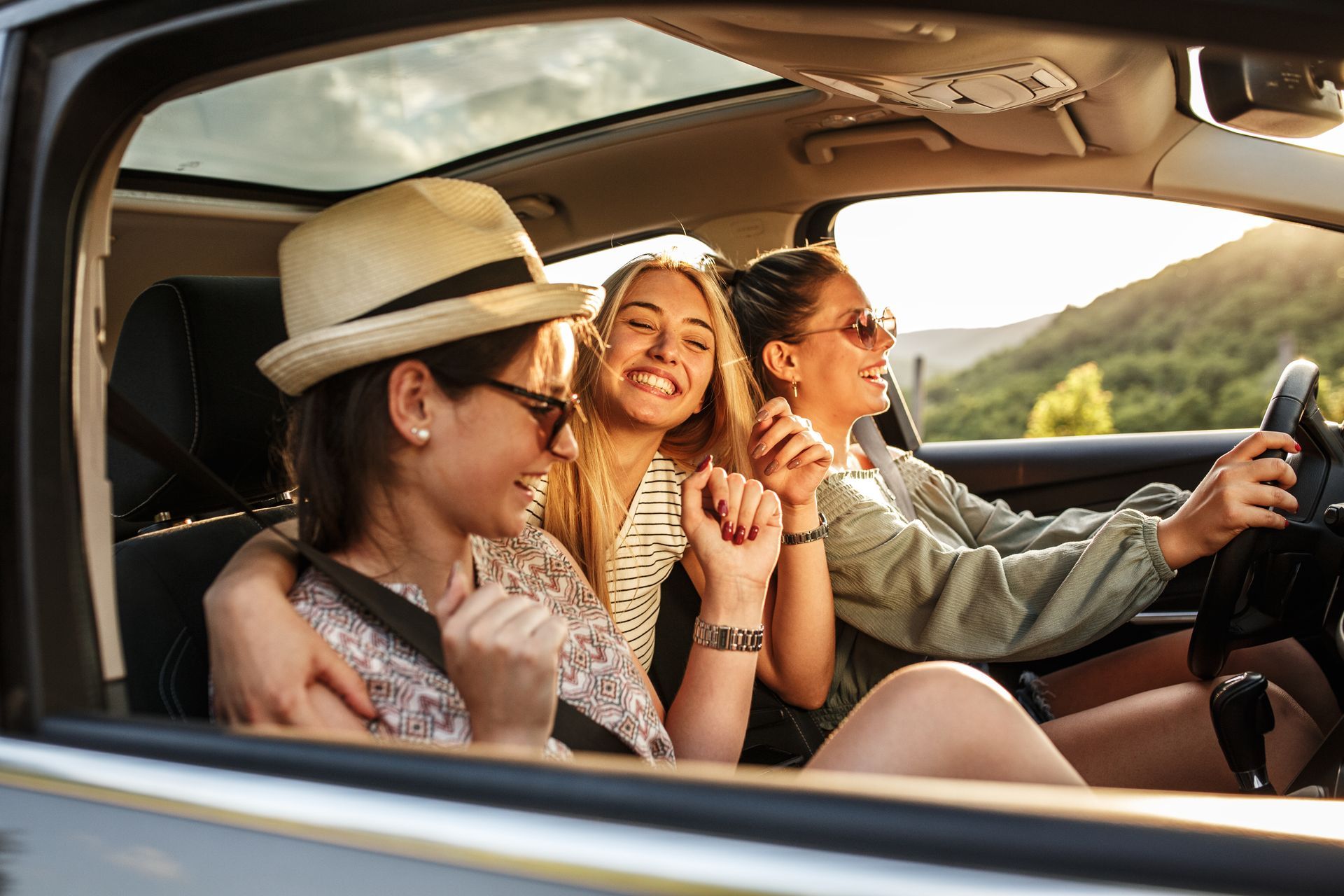 Three women smiling in a car, one driving, scenic background, sunlight.