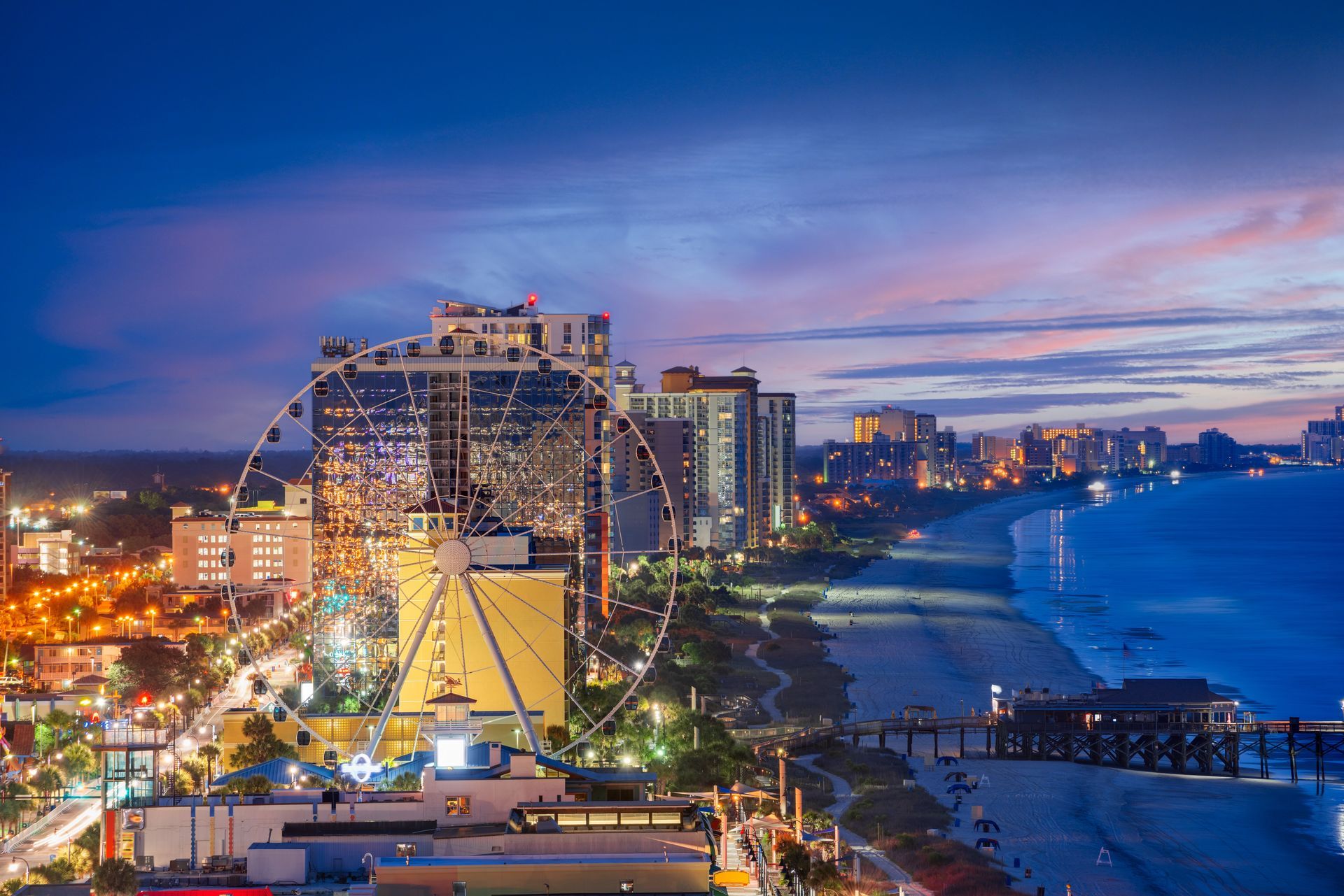 Myrtle Beach at twilight, featuring the SkyWheel, hotel skyline, and a fishing pier extending into the ocean.