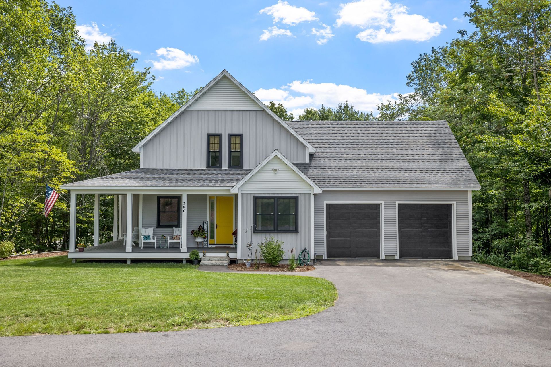 A gray two-story house with a wrap-around porch, yellow front door, and two-car garage, surrounded by green trees.