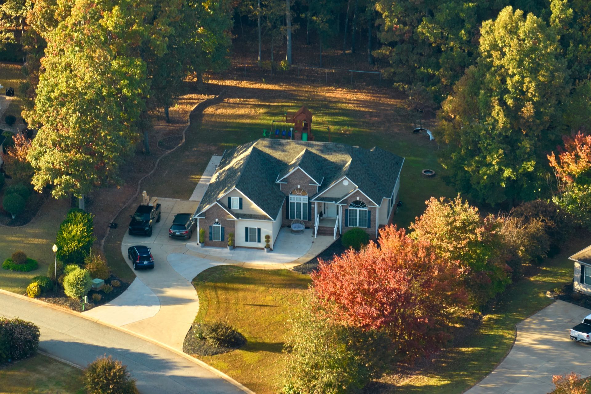 An aerial view of a suburban brick house with a concrete driveway and surrounding trees during the autumn season.