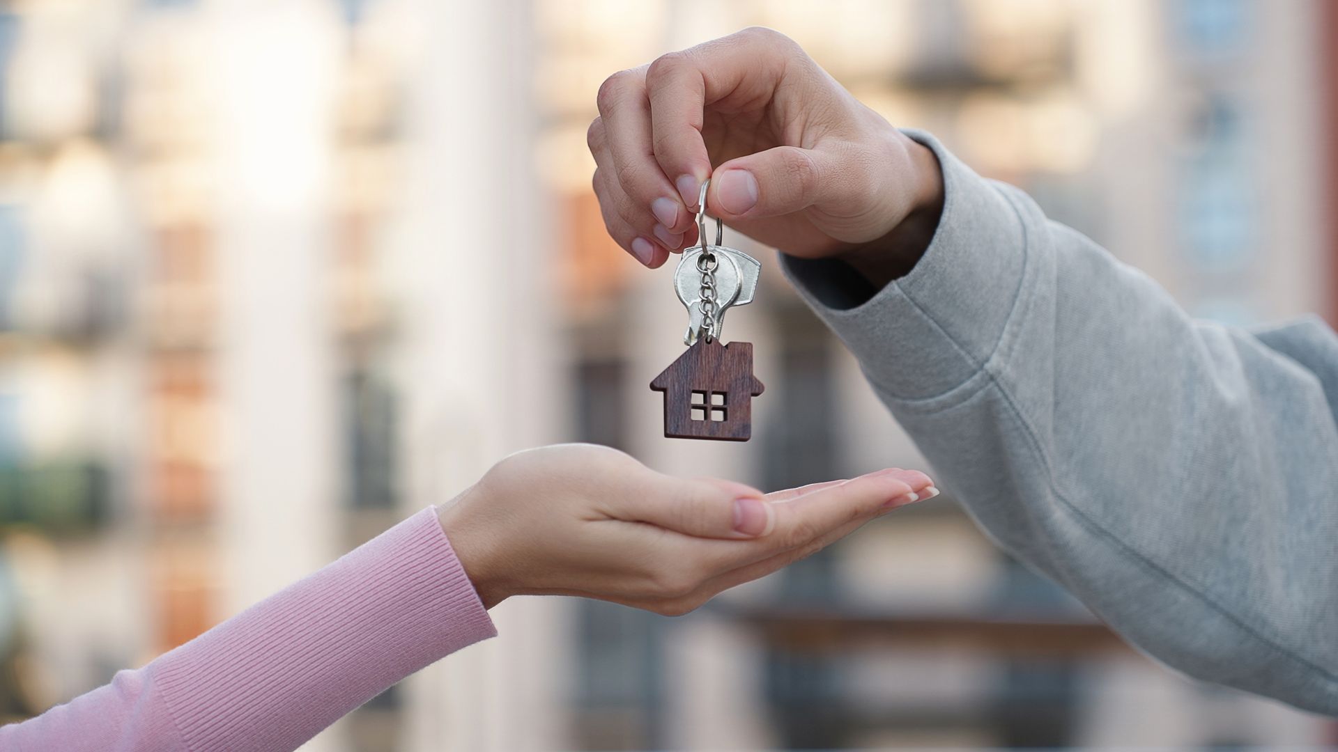 One person hands a key with a small house-shaped keychain to another person against a blurred background.