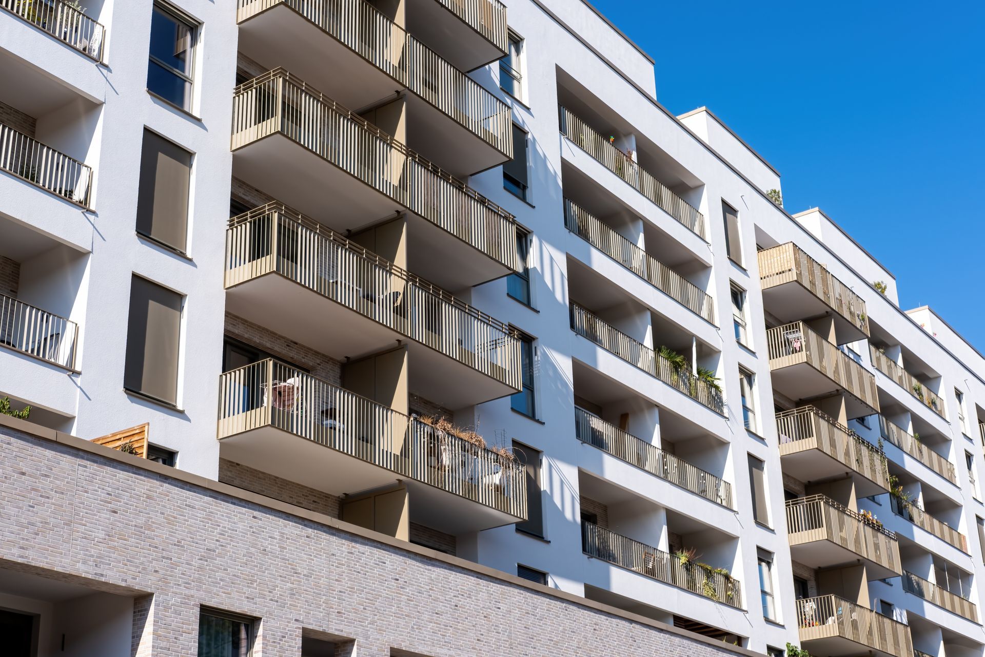A modern, multi-story white apartment building with stacked balconies and metal railings against a clear blue sky.