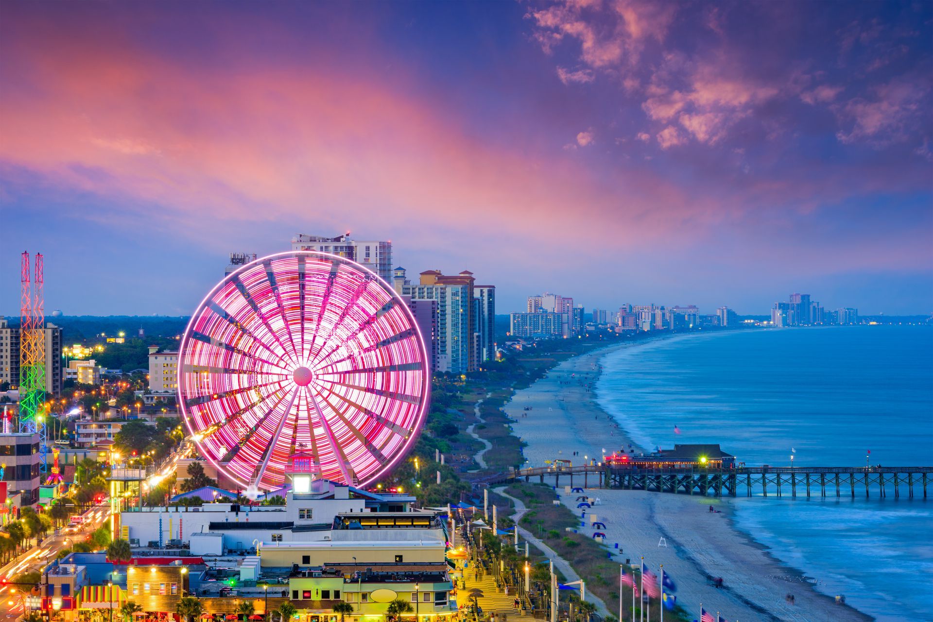 Ferris wheel on a beach at sunset; colorful sky, ocean, and city skyline.