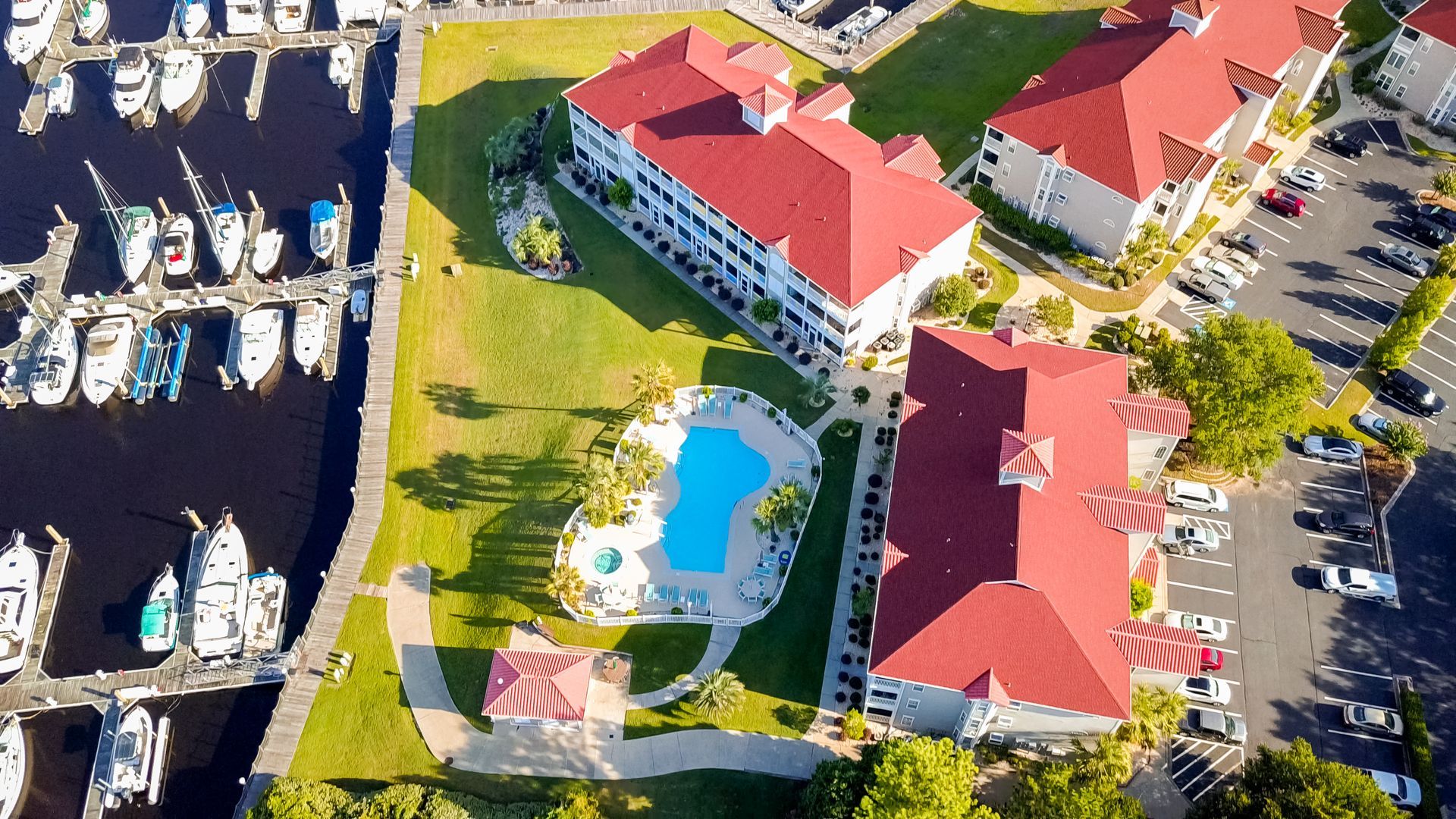 Aerial view of buildings with red roofs, a pool, marina, and parked cars.