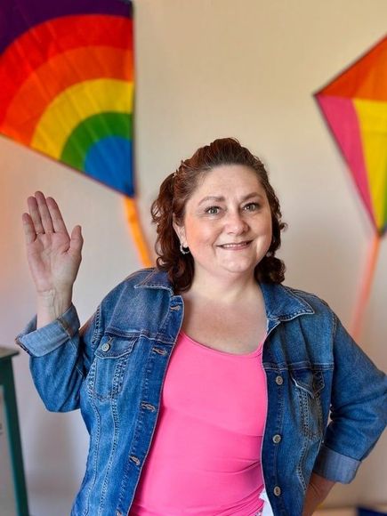 A woman in a pink tank top and a denim jacket is waving in front of a rainbow and kites.