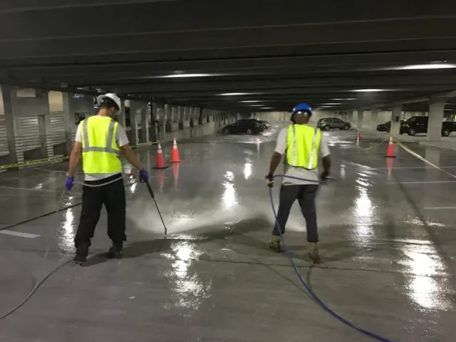 Two workers power washing a concrete parking garage floor, wearing safety vests and helmets.