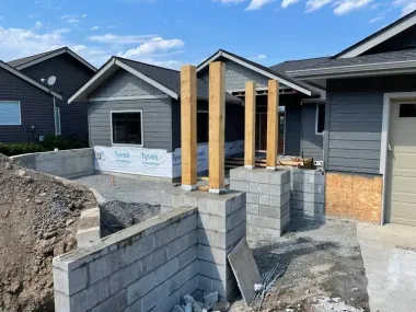 Construction site: Exterior of a gray house with wooden support beams on concrete blocks; blue sky.