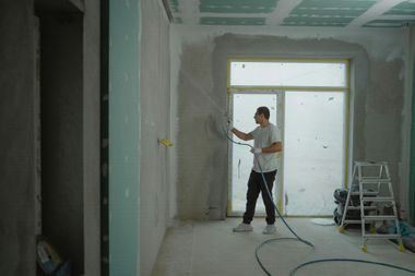Man spraying wall with paint in a room under construction, with a window and ladder.
