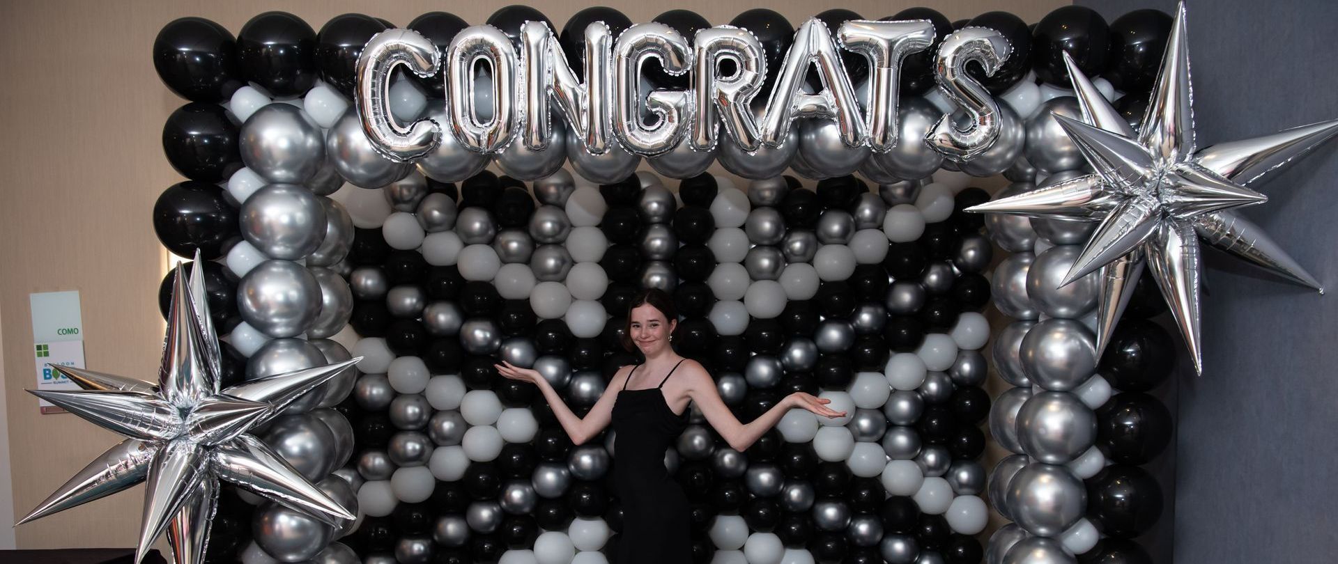A woman in a black dress stands in front of a balloon arch that says