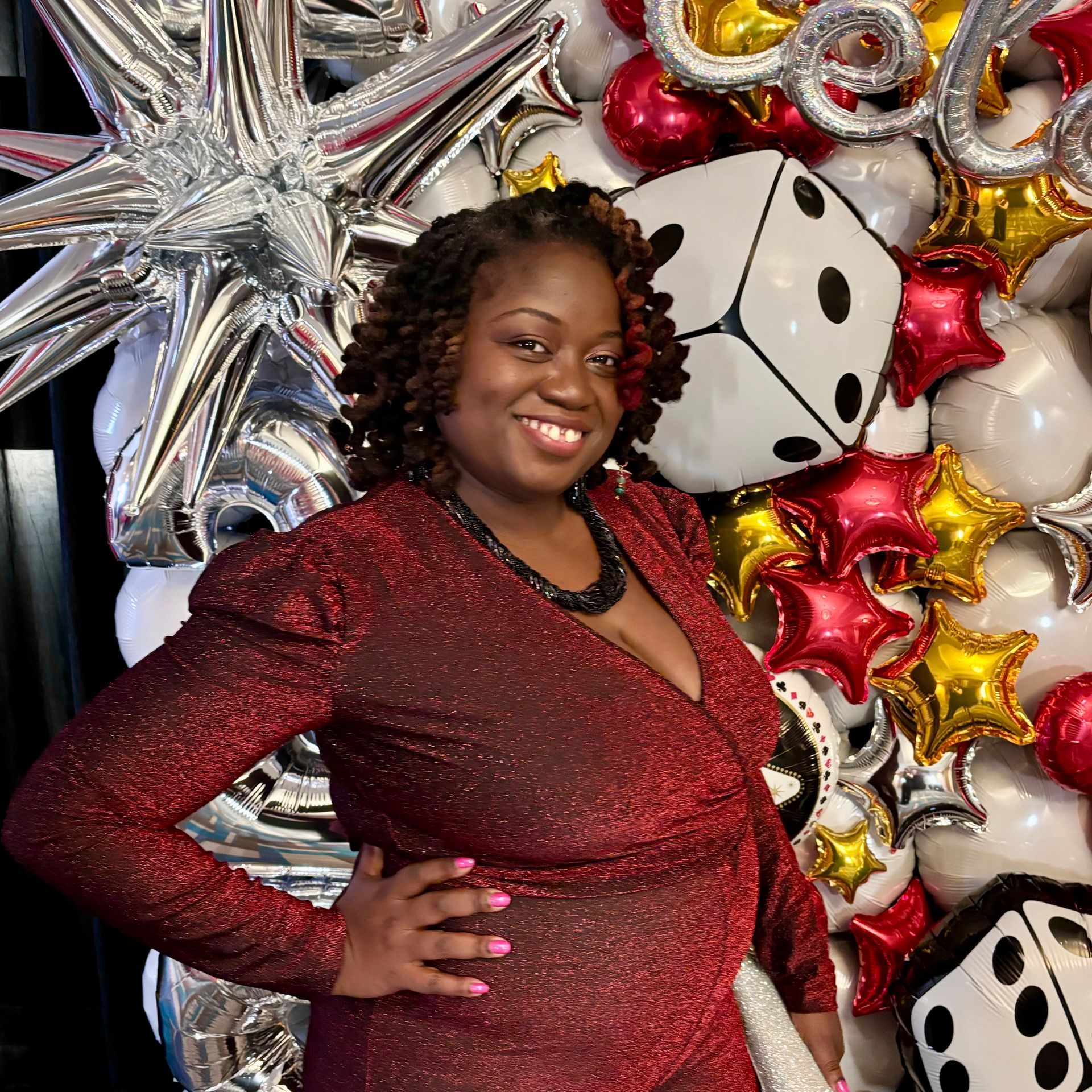 Woman in red dress smiles, posing by dice and star balloons.