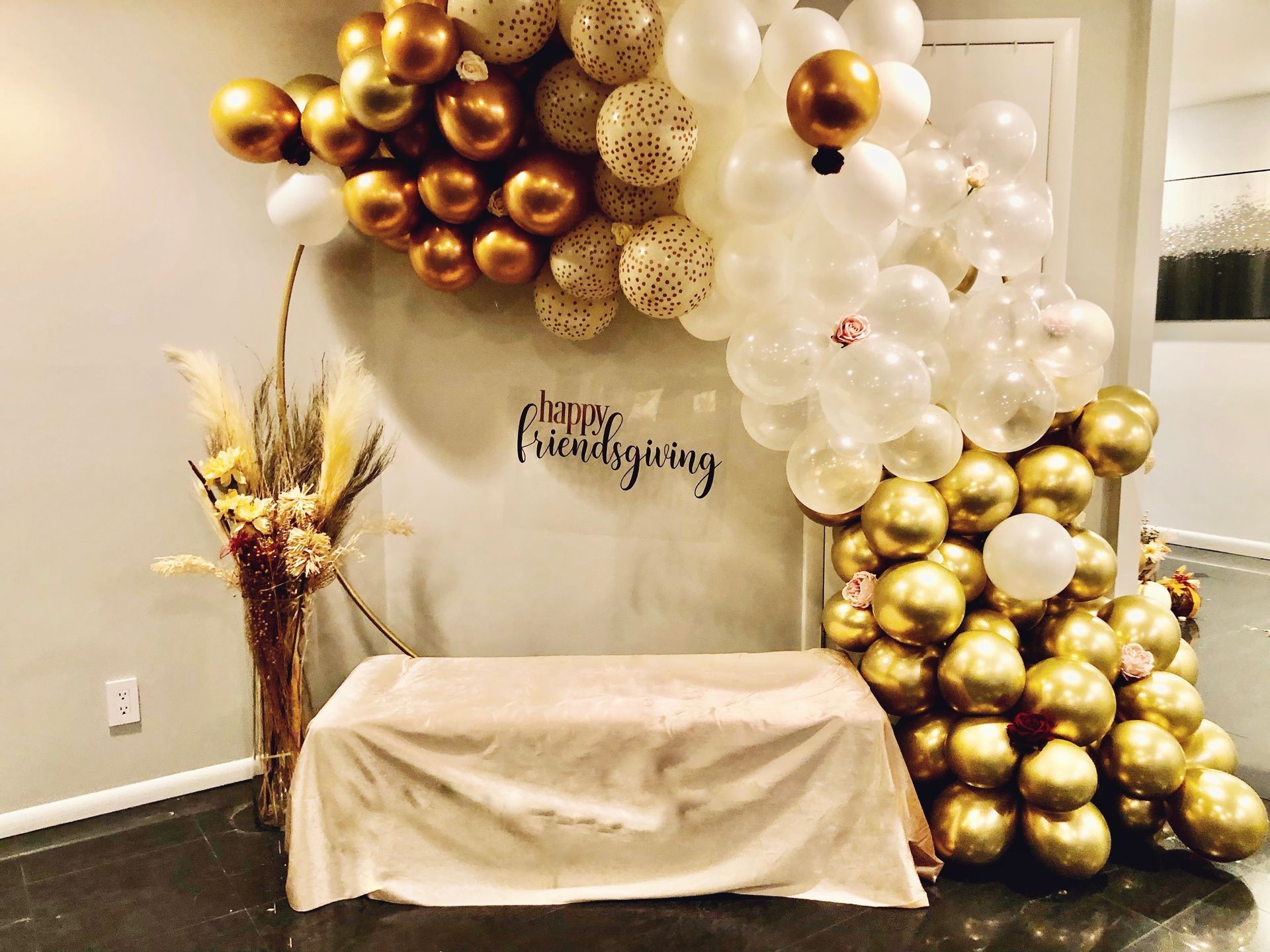 Gold and white balloon arch over a table with a beige cloth, against a wall. Includes dried flowers and text.