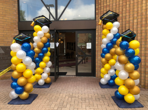 Four balloon columns in graduation colors flank a doorway. Each column has a black mortarboard hat on top.