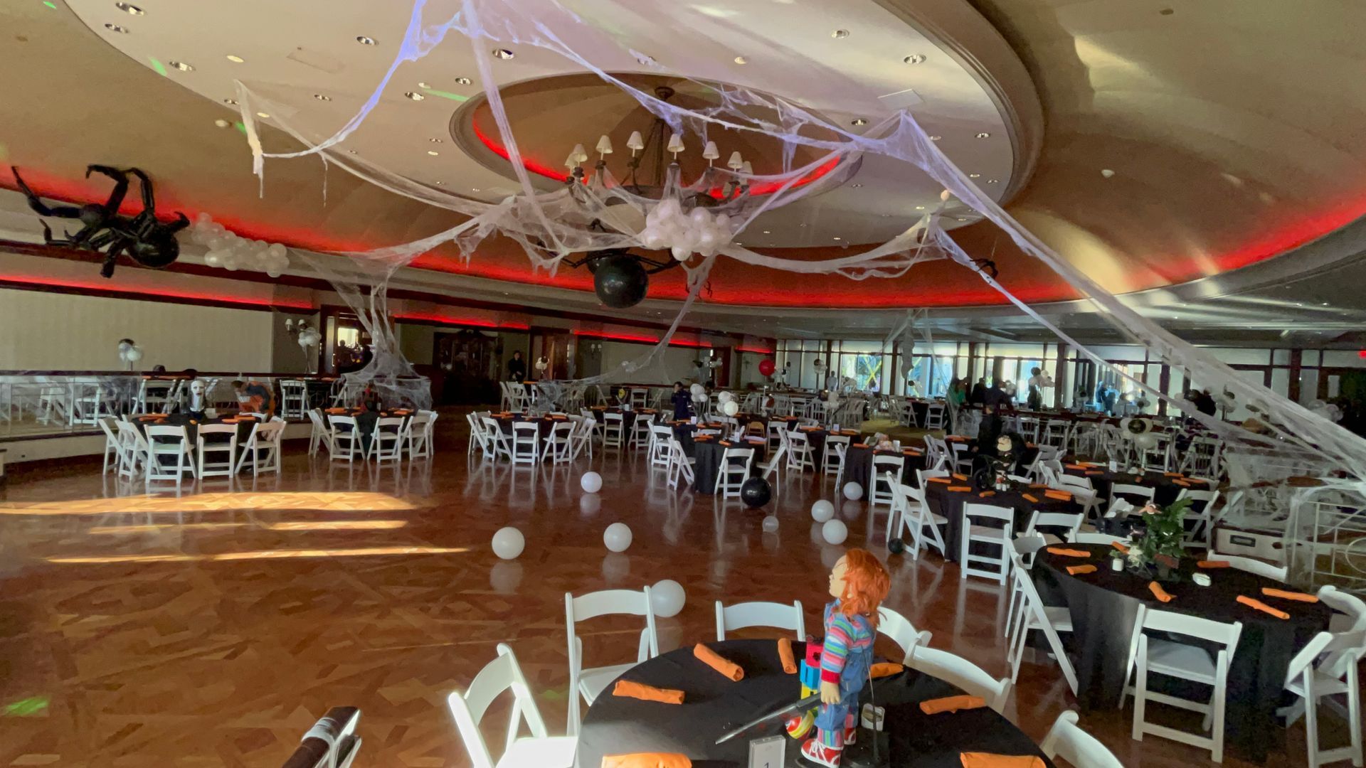 Halloween-decorated event space with tables, chairs, fake spiderwebs, and a Chucky doll centerpiece.