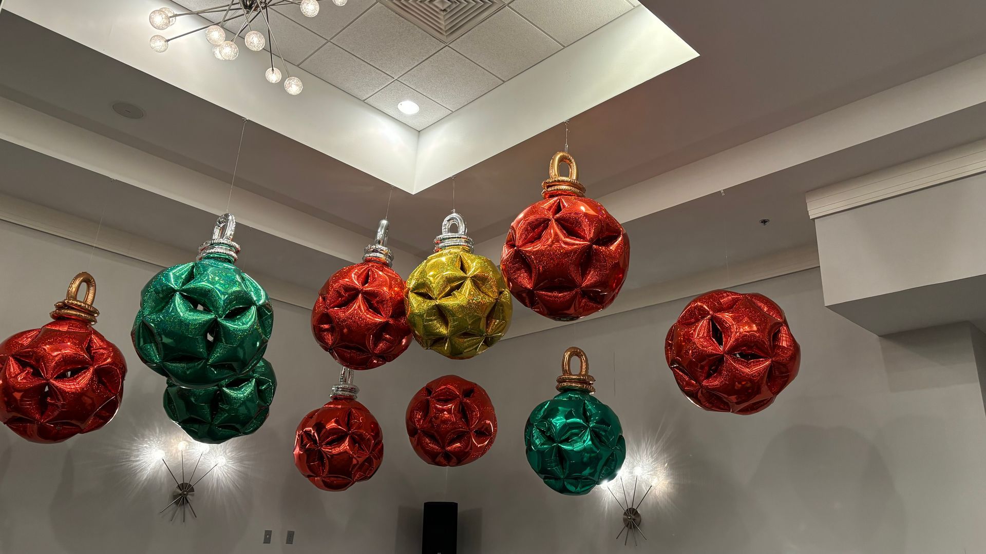Giant, colorful, glittery ornaments hanging from a ceiling. Red, green, and gold baubles decorate a room.