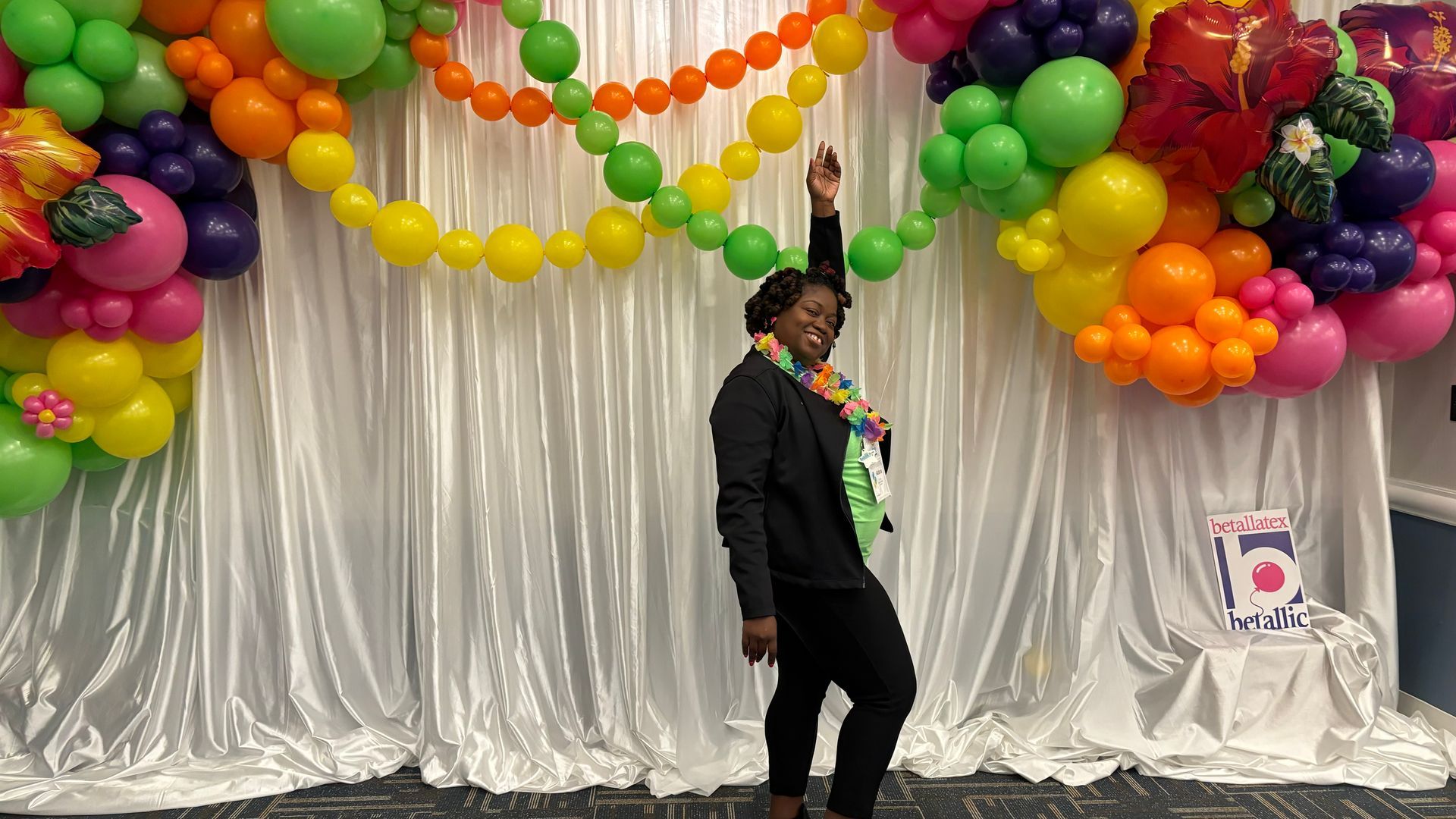 Woman smiling, posing with arm raised. Balloons in rainbow colors decorate the backdrop.