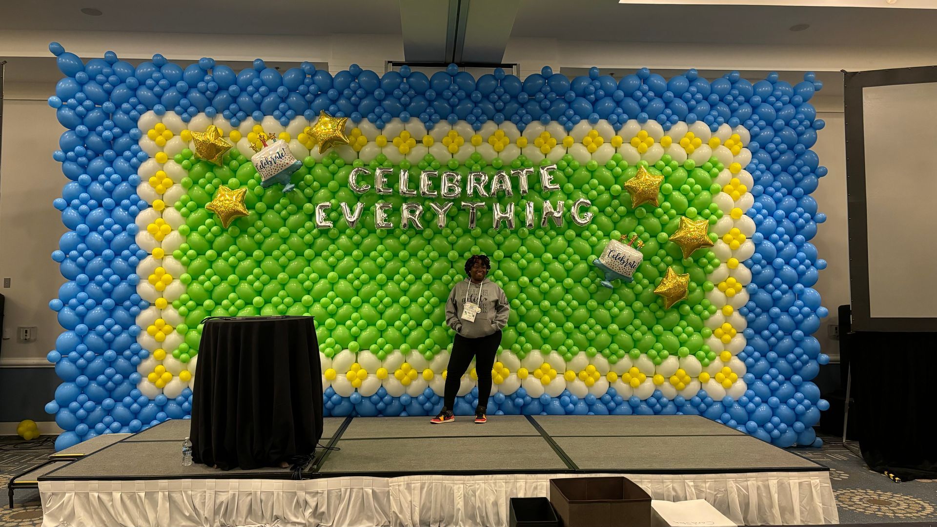 Woman standing on a stage in front of a balloon backdrop that says 
