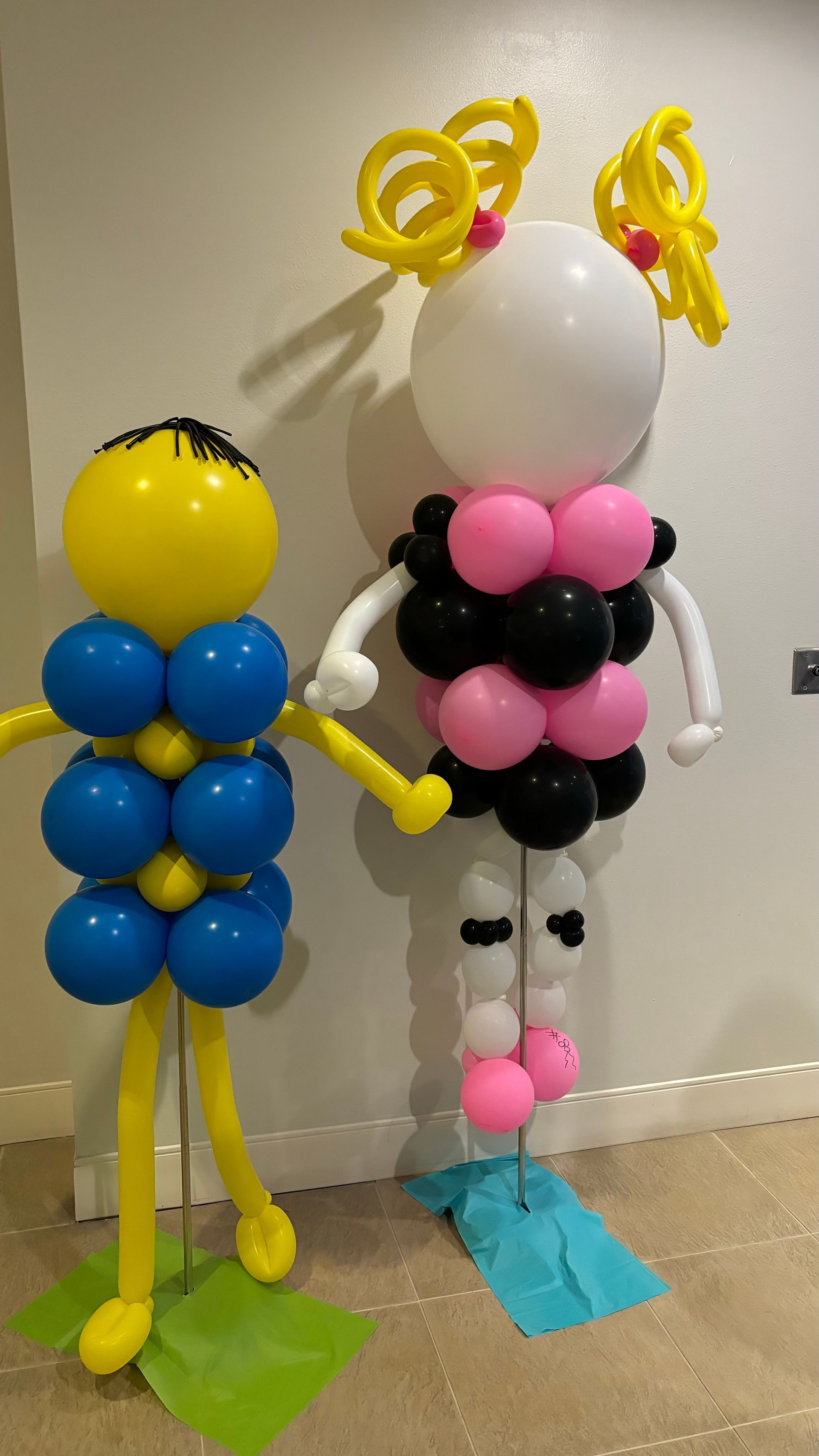 Two balloon sculptures: yellow and blue figure holding hands with a white and black figure, indoors.